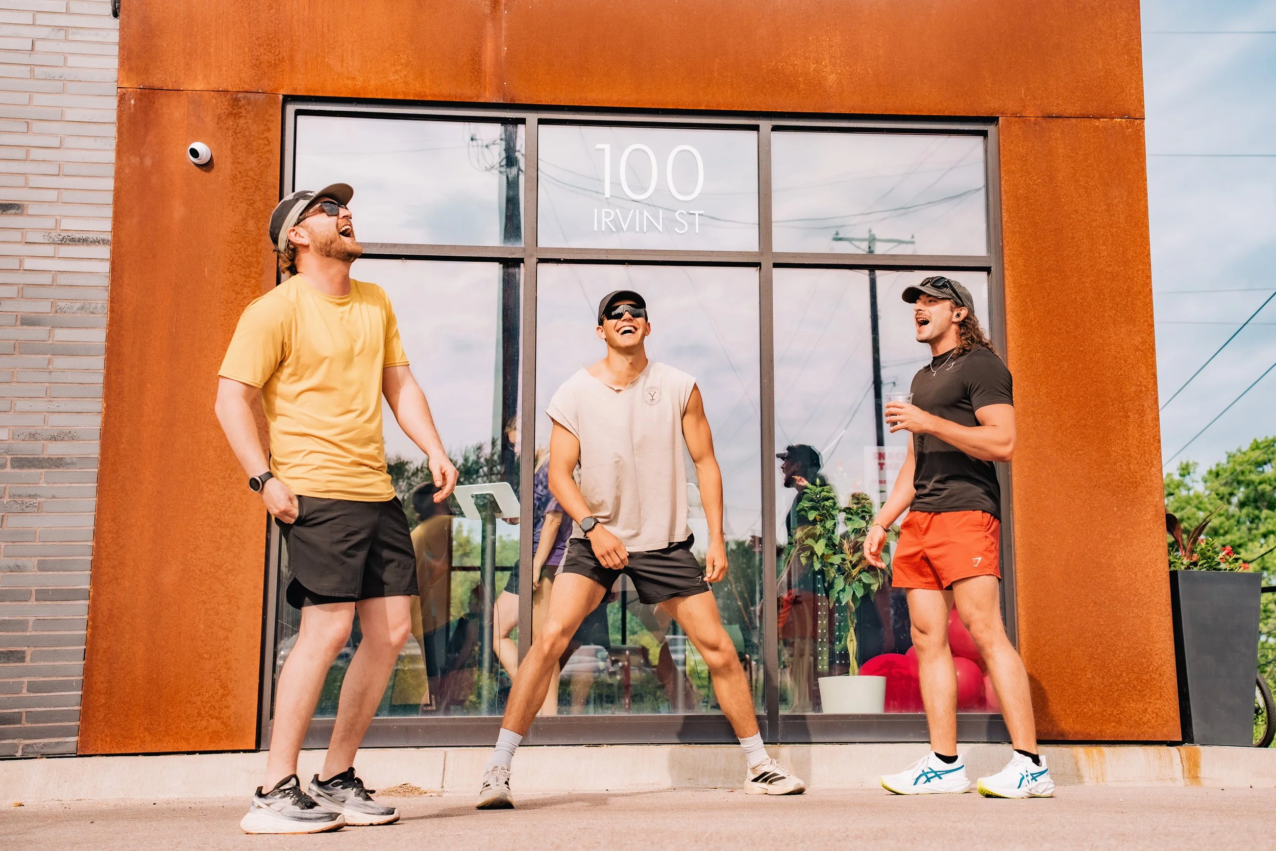Three young men standing outside a modern building labeled '100 IRVIN ST,' laughing and posing casually in athletic wear and sunglasses.