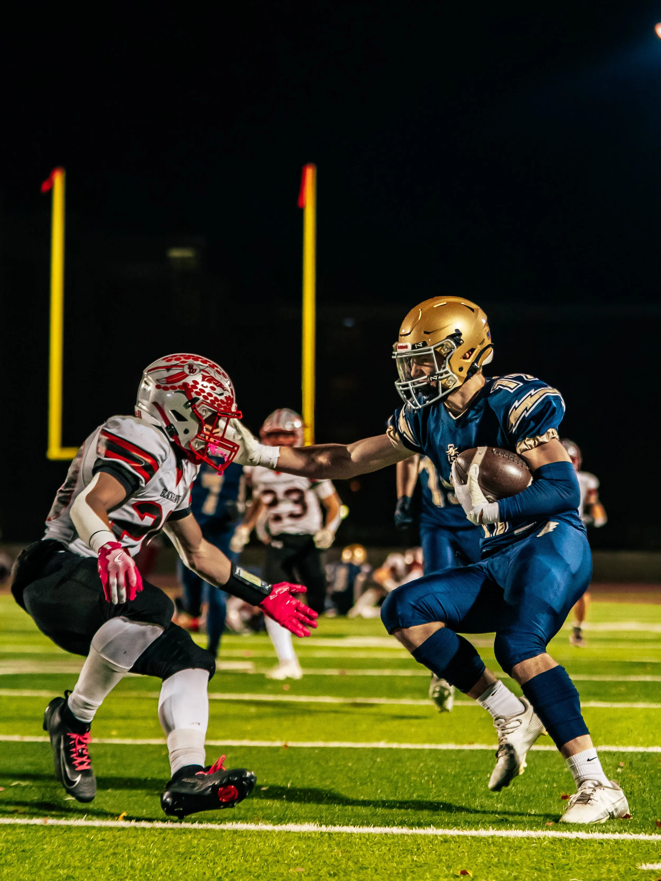 A nighttime American football game with players in blue and white uniforms. One player in blue is holding a football and attempting to run past a player in white and red who is reaching out to block him. The game is on a brightly lit field with yello