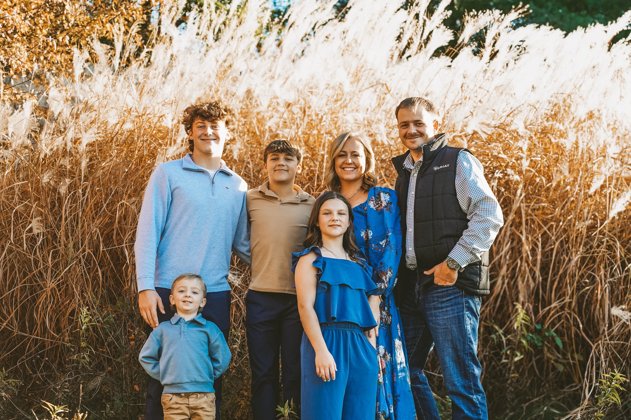 A family of seven standing outdoors in front of tall, dry grass and trees during autumn, smiling at the camera.