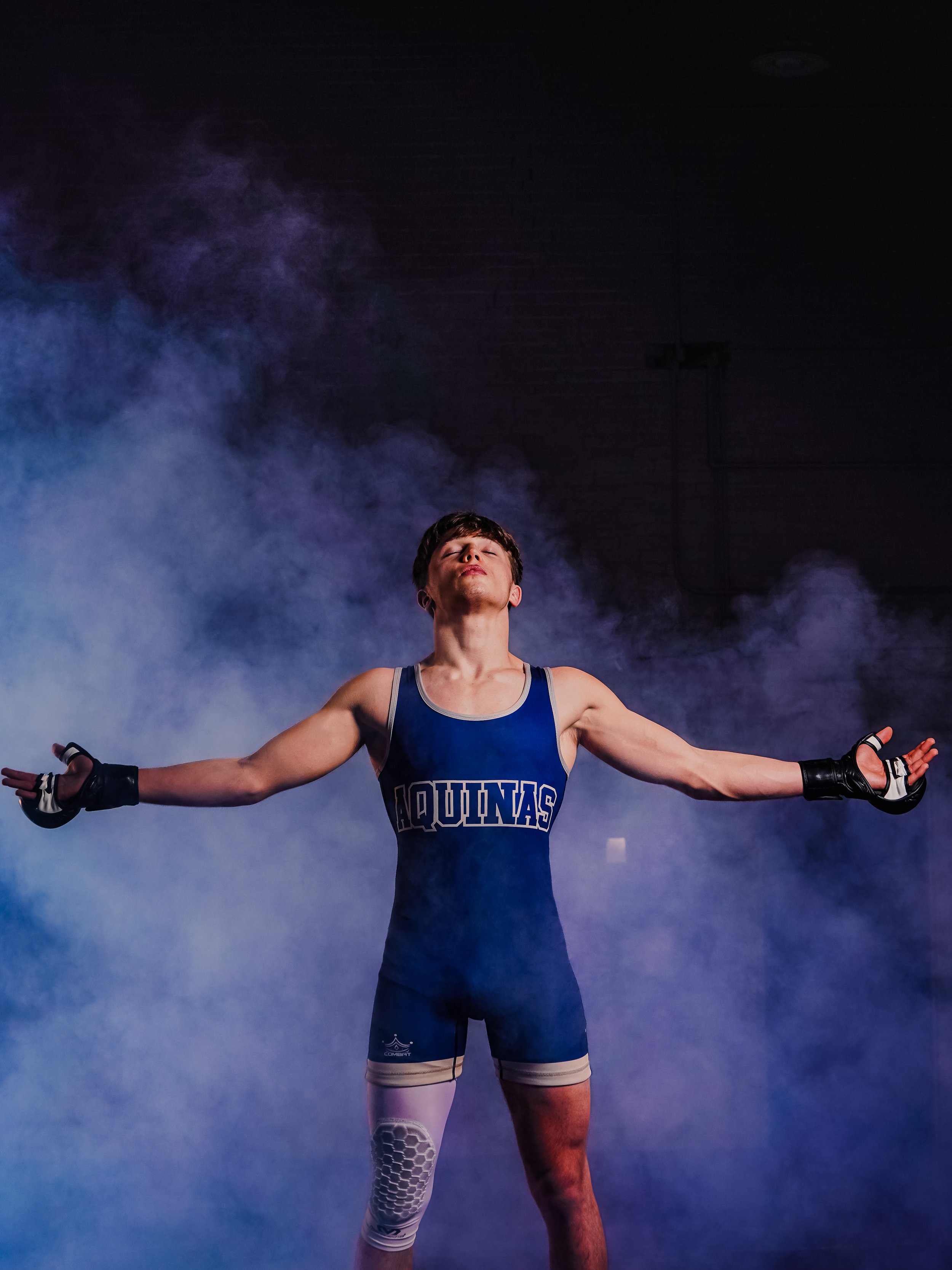 A male athlete with short dark hair is standing with his arms outstretched, eyes closed, wearing a blue and white sports uniform with 'AQUINAS' written on the front, black gloves, white athletic gear on one leg, and a white knee brace, surrounded by 