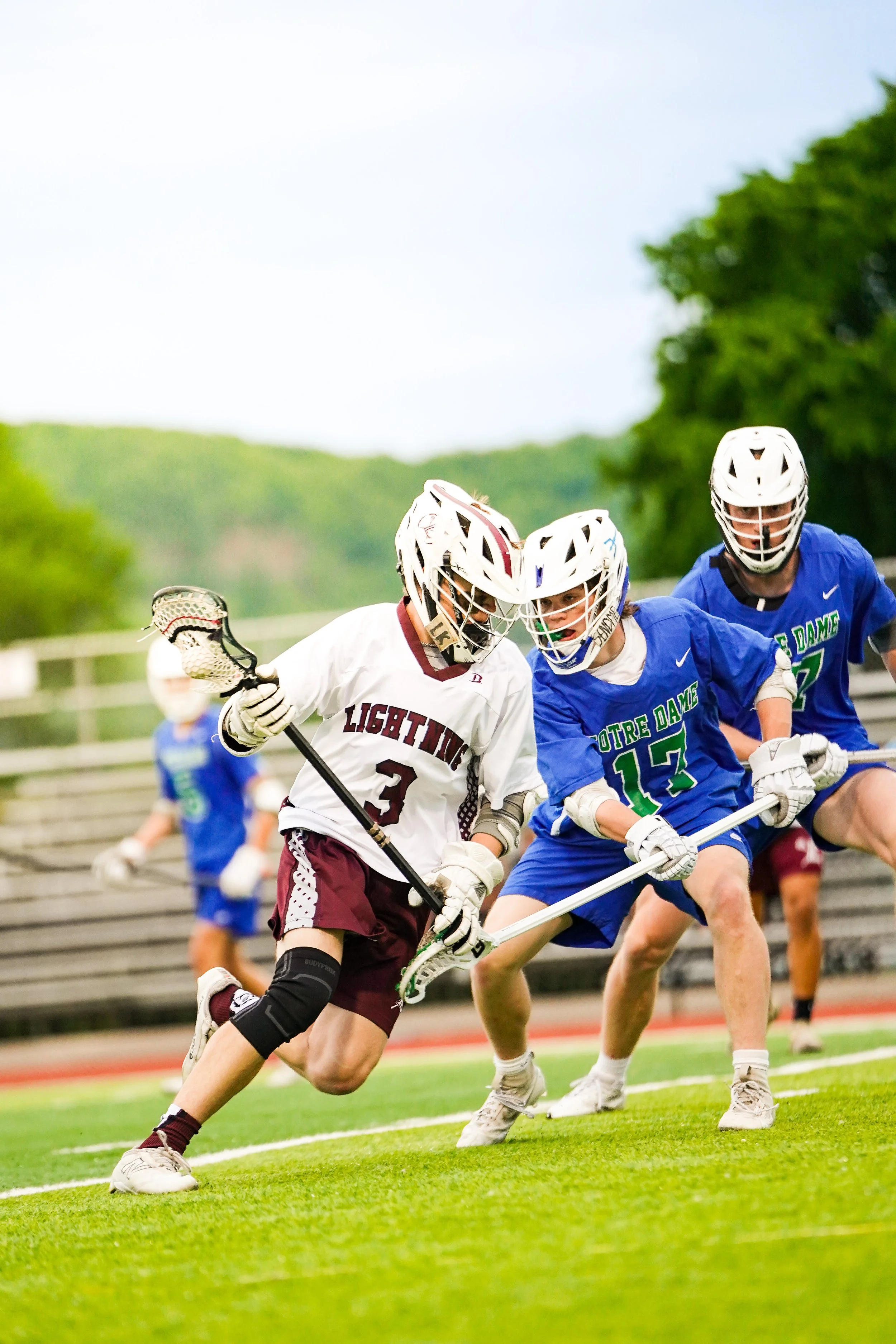 High school lacrosse game with players in white and blue uniforms competing on the field.