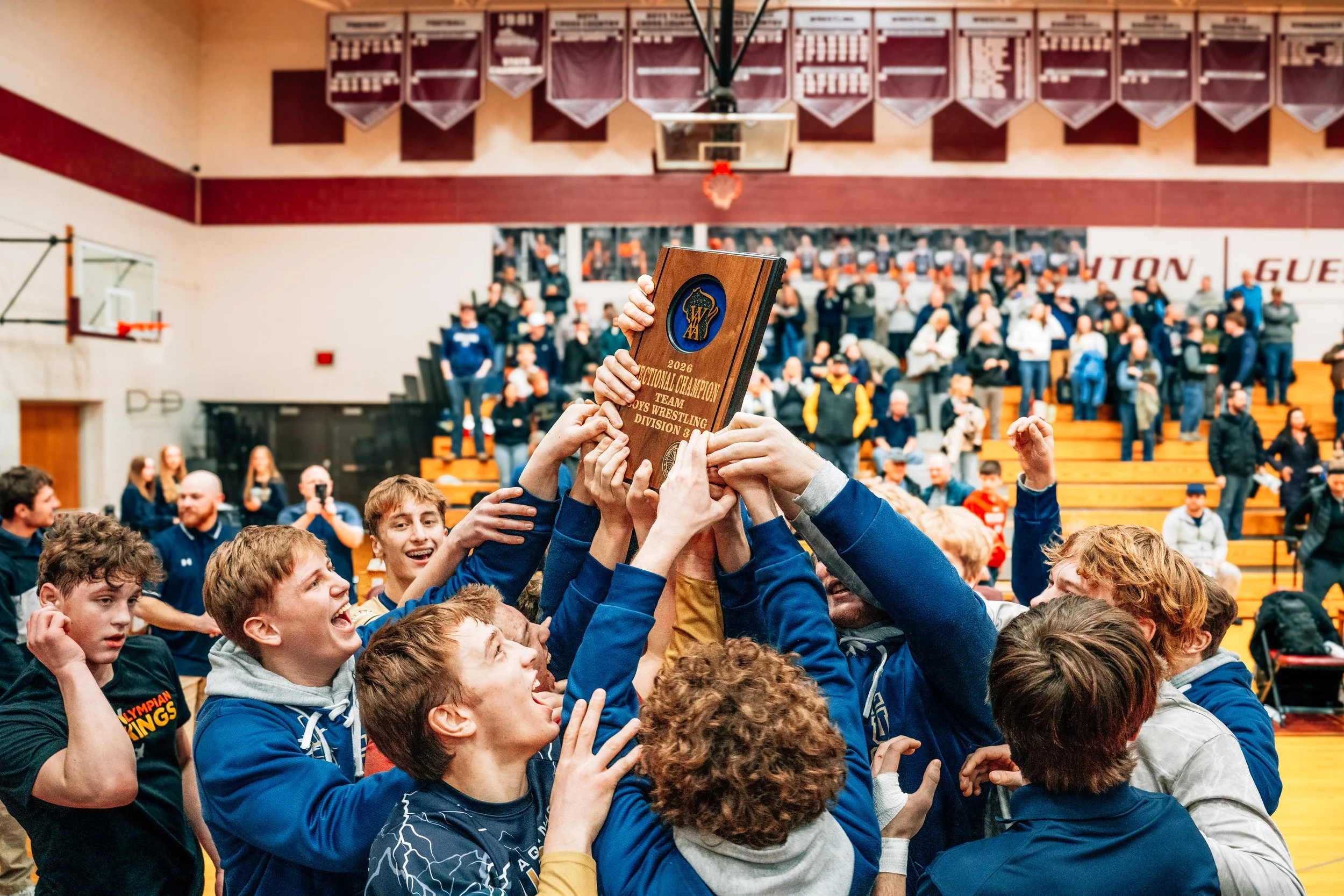 A group of young people celebrating as they lift a wooden trophy with a blue emblem and the text '2026' in a gymnasium. The background shows a basketball hoop, banners, and spectators.