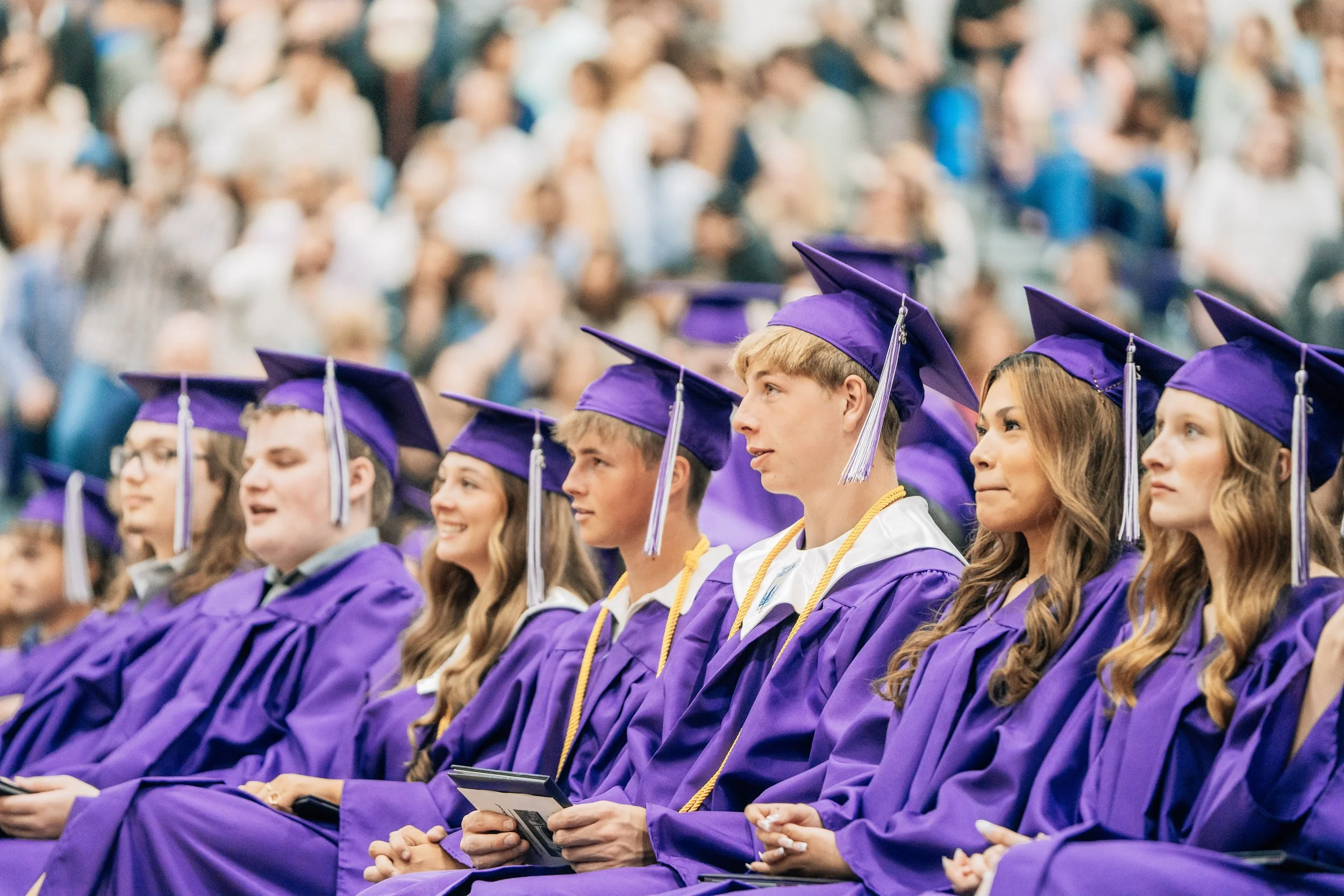Students in purple graduation caps and gowns sitting in an indoor auditorium.