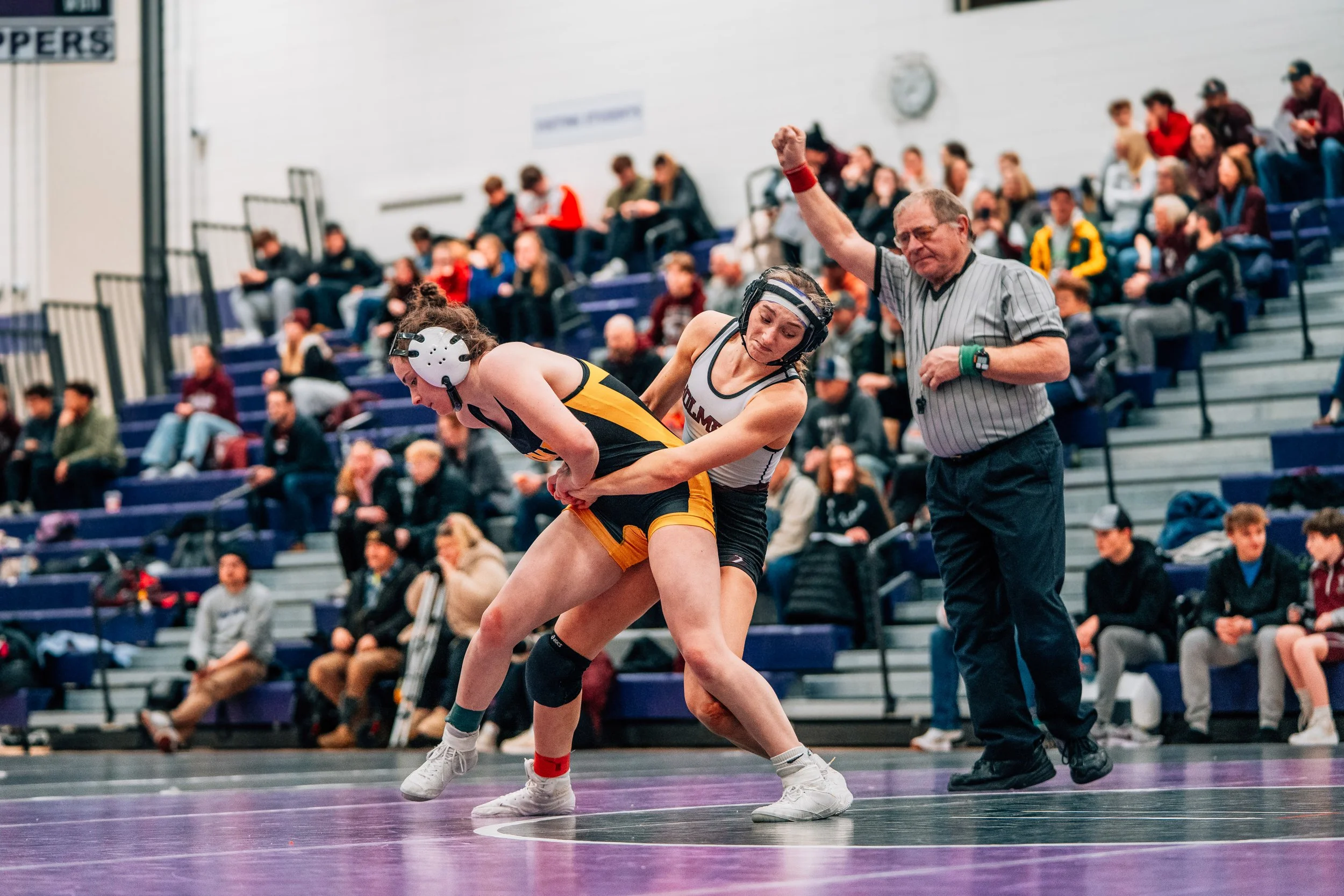 Two female wrestlers compete on a wrestling mat, one is in a black and yellow uniform and the other in maroon and white. An official in a striped shirt is officiating the match, raising his right arm. Spectators sit on bleachers in the background wat