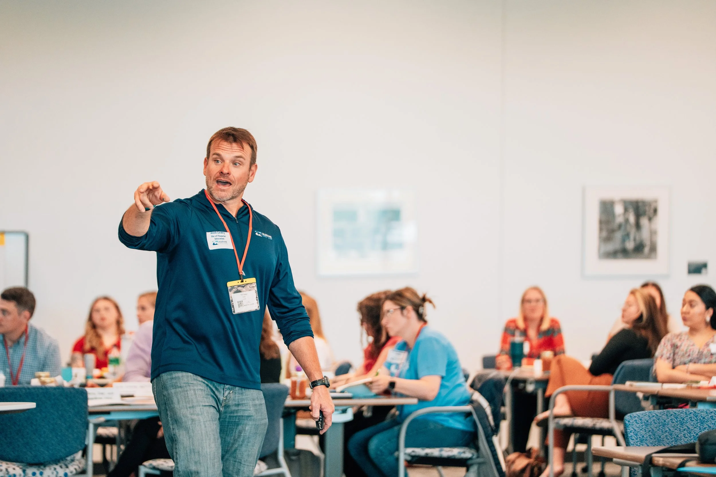 A man with a microphone in his hand standing at the front of a conference room, speaking to an audience of seated people. The audience is a mix of men and women, some taking notes, with a few looking at the speaker attentively. The room has bright li