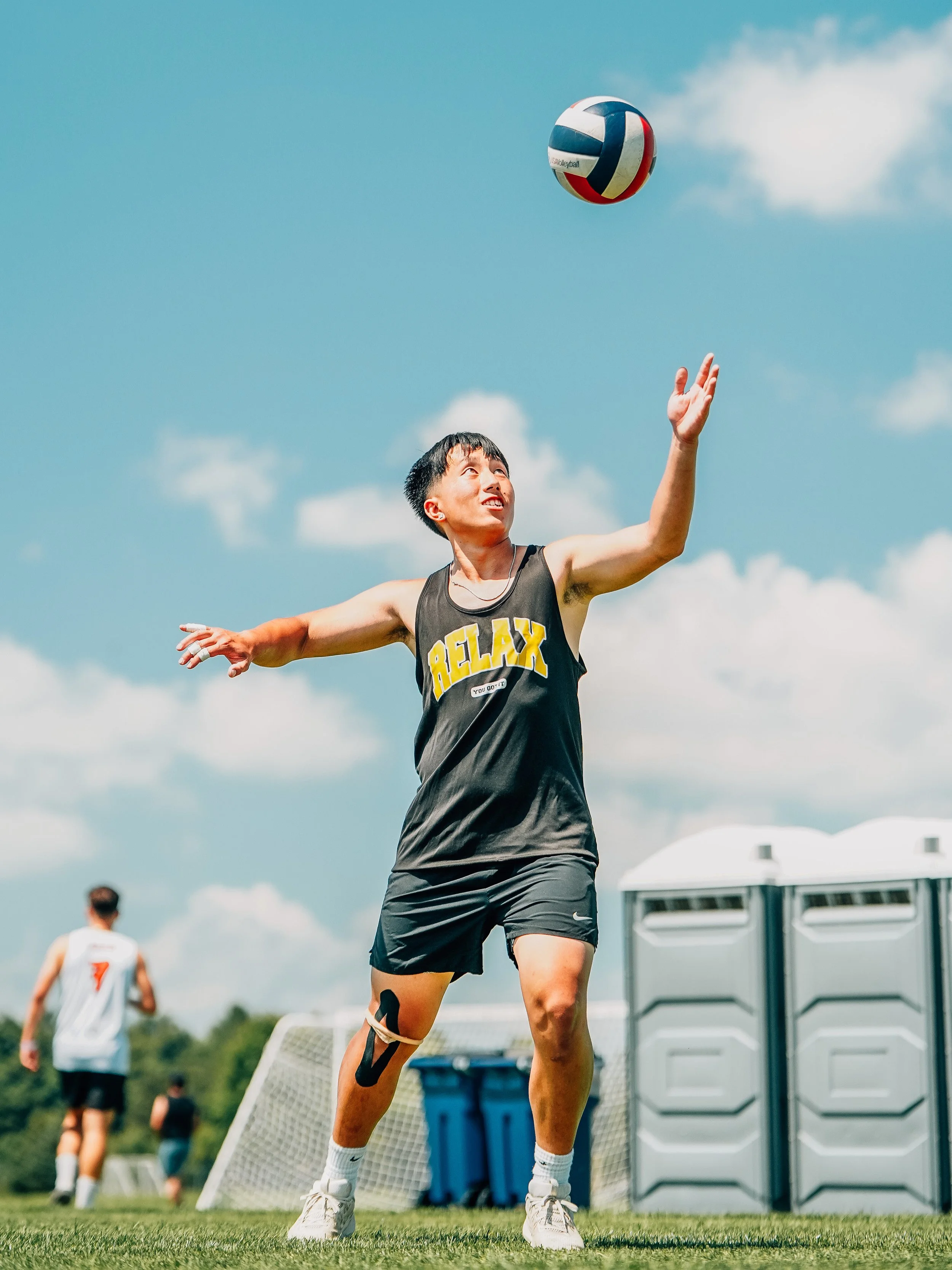 A young man playing volleyball on a grassy outdoor field, with a large portable storage container behind him, blue sky with some clouds overhead, and people in sports attire in the background.