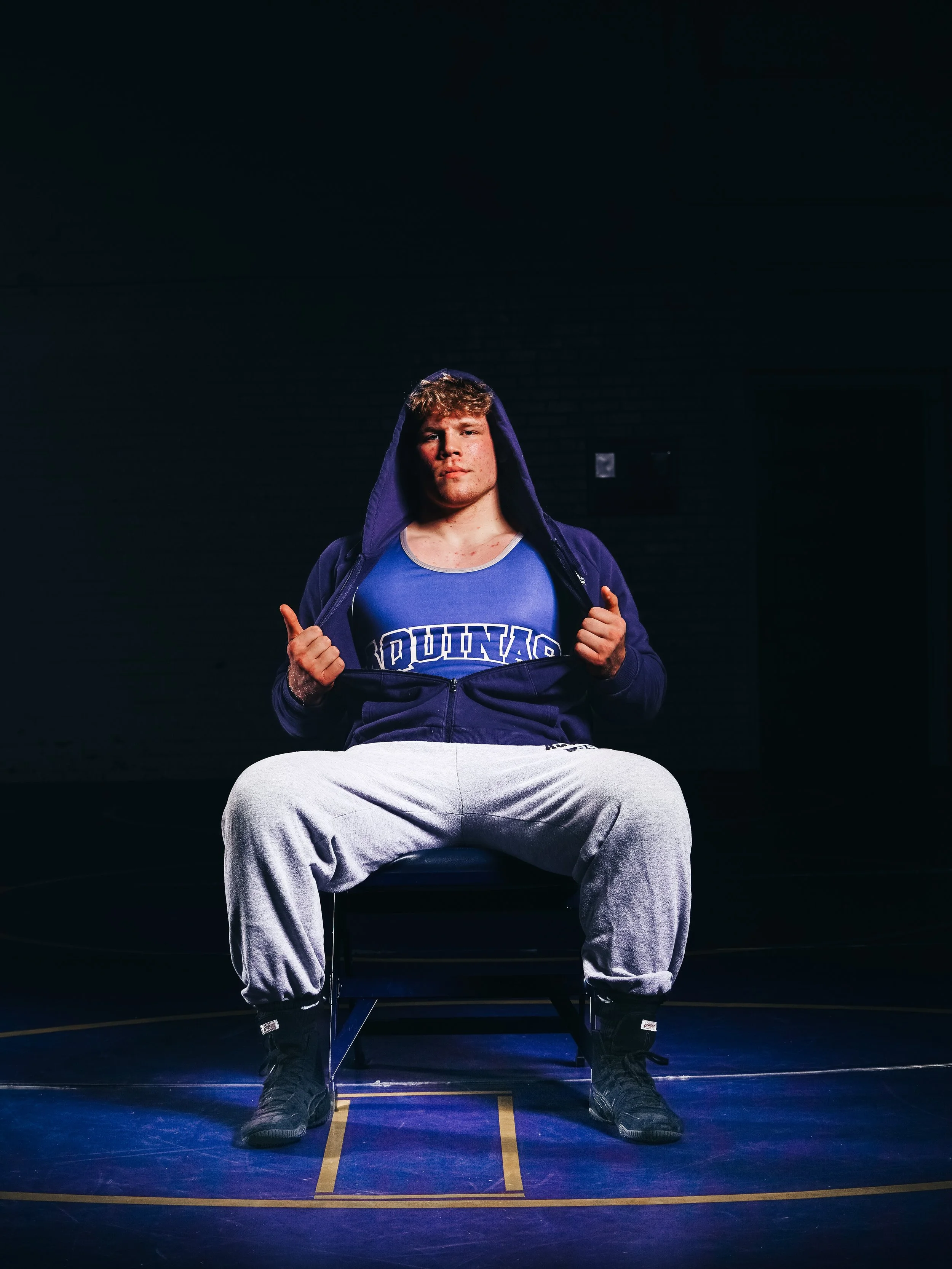 Young male athlete with curly hair, wearing a blue sports shirt reading "DUNDAS" and a dark hoodie, seated on a chair in a dimly lit gymnasium with a dark background and blue lighting, holding the edges of his hoodie.
