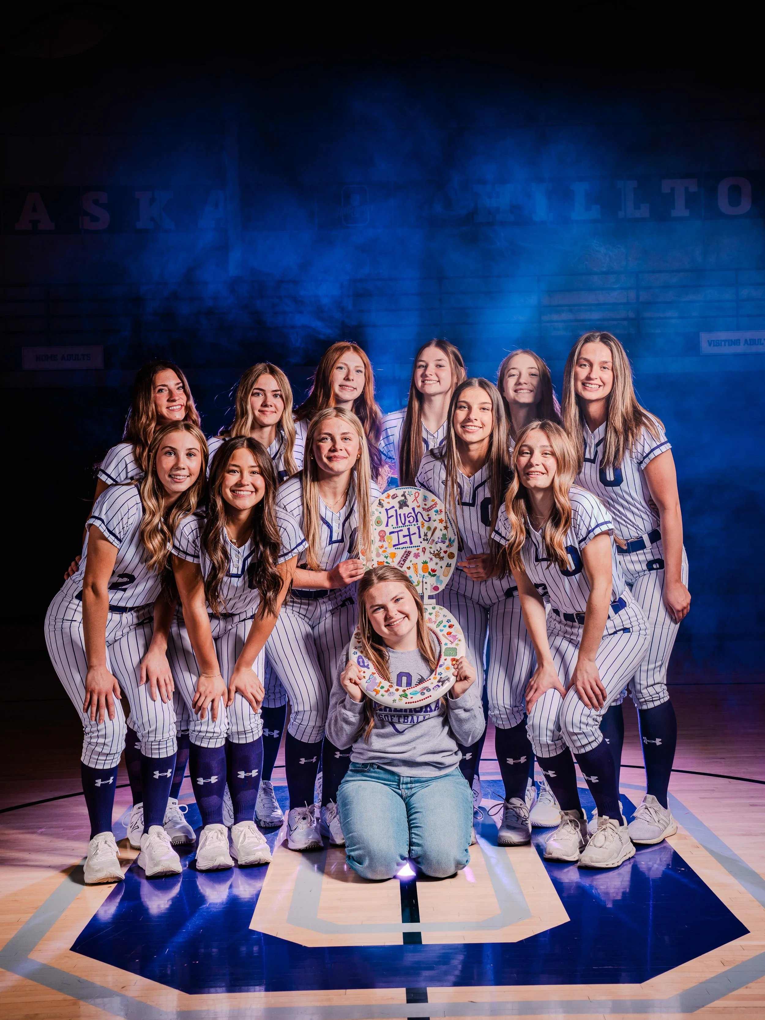 A group of girls in softball uniforms posing on a basketball court, one girl sitting on the floor holding a decorated softball wreath and a balloon with 'Flip' written on it.