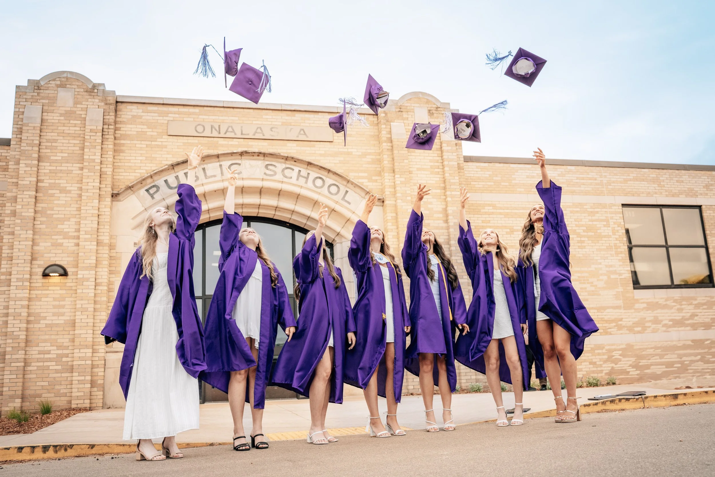 High school graduates in purple gowns toss caps into the air in front of a brick school building.