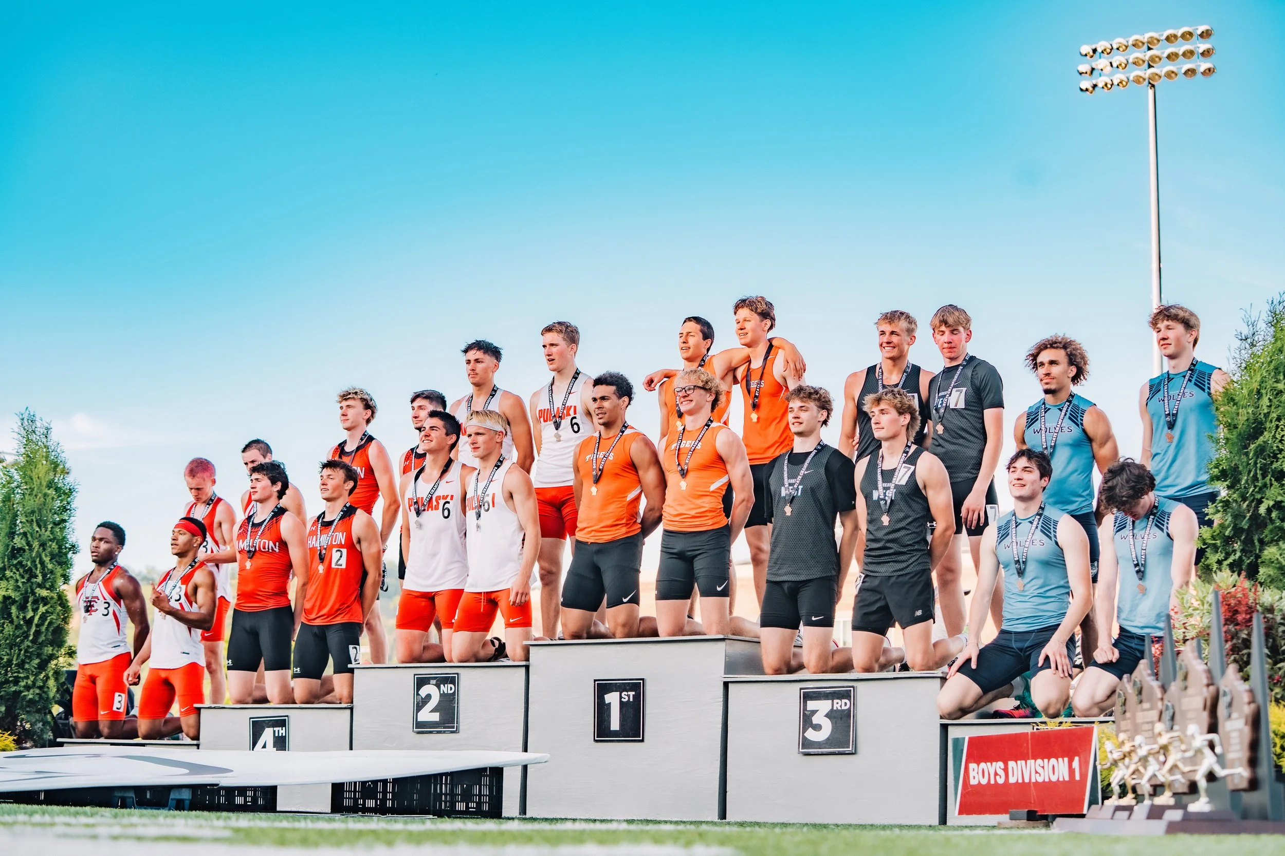 Young male athletes standing on a winners' podium after a race, wearing medals, with first, second, and third-place signs, under a clear blue sky.