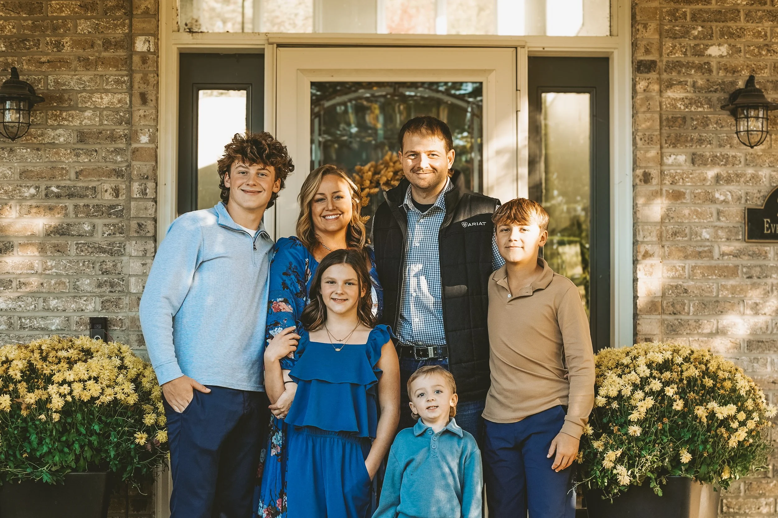 A family of seven people standing on front porch of house, smiling, surrounded by yellow flowers.
