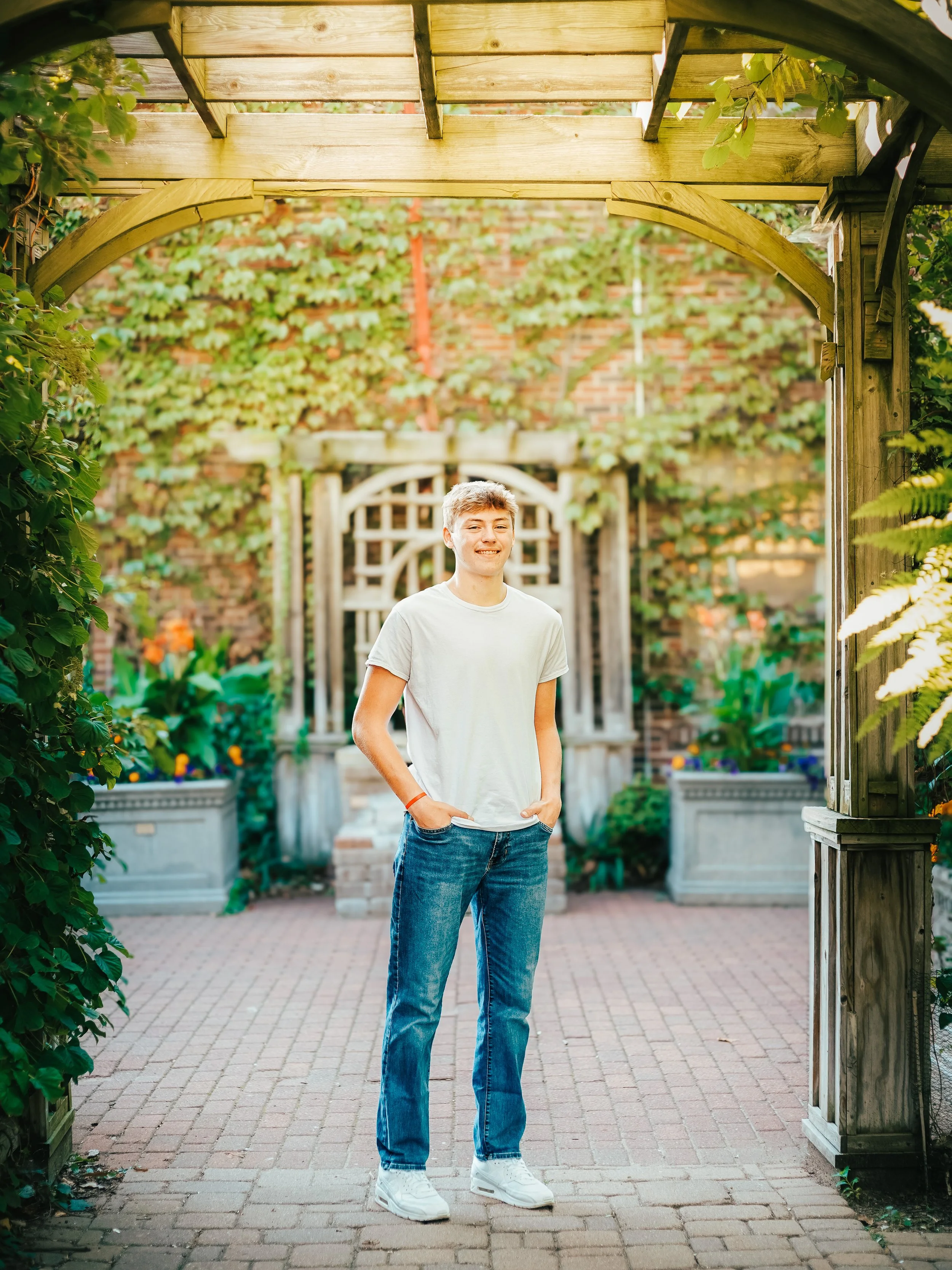 A young man with light brown hair, wearing a white t-shirt and blue jeans, standing on a brick patio under a wooden garden archway, smiling and looking at the camera, with greenery and a lattice fence in the background.
