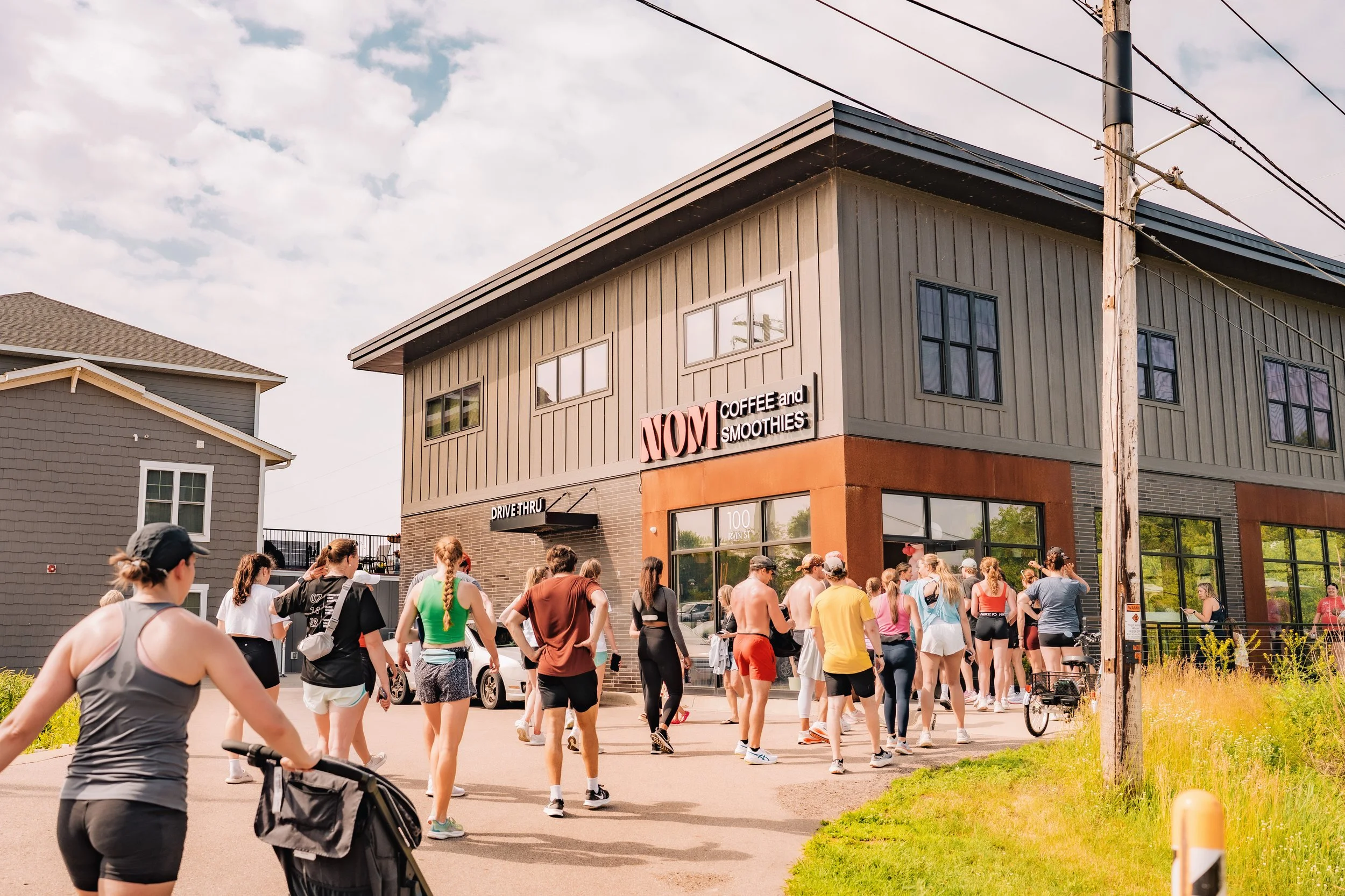 A large group of people in athletic clothing gathered outside a building with a sign that reads 'NOM Coffee and Smoothies' during daytime with partly cloudy skies.