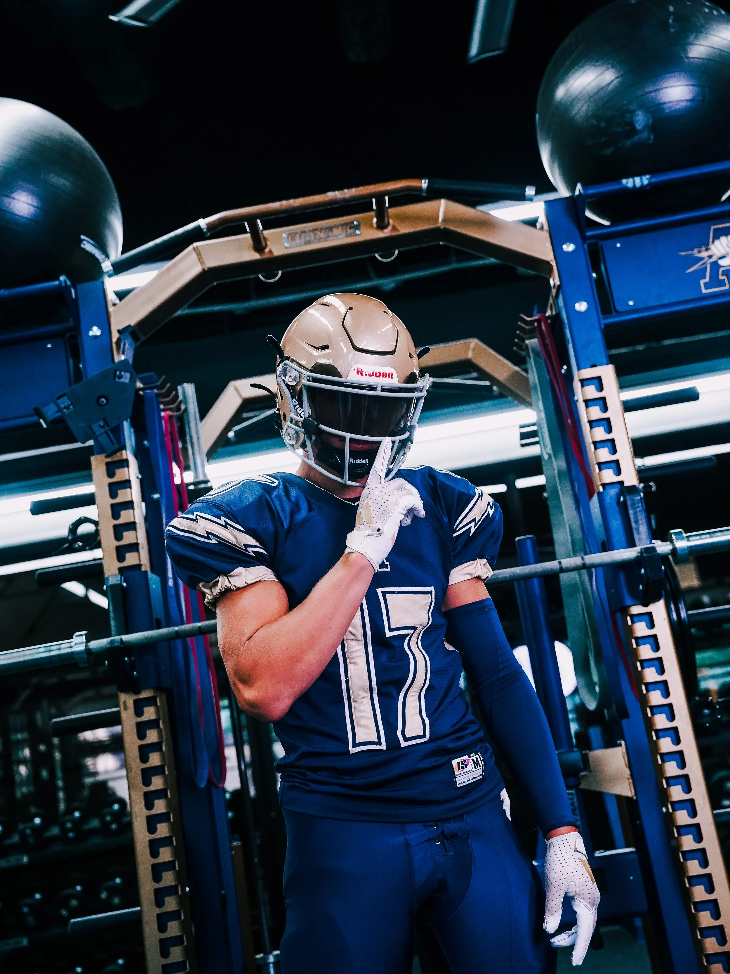 A football player standing in a gym, wearing a blue jersey with the number 7, a beige helmet with a face guard, and white gloves. The player is holding a finger to his lips in a 'shh' gesture, with gym equipment and weights visible in the background.