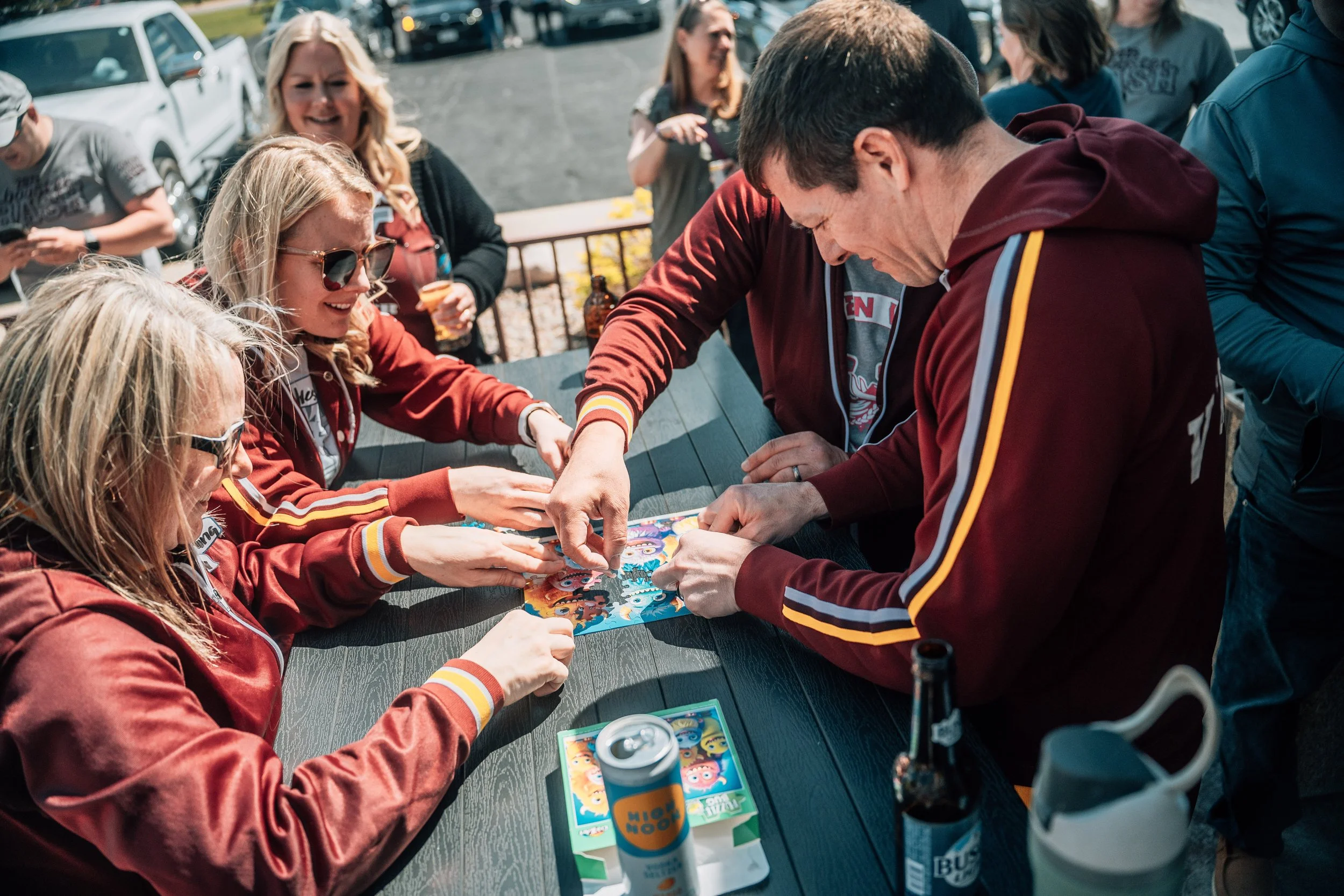 People gathered outside around a table playing a board game on a sunny day, with cars parked in the background.