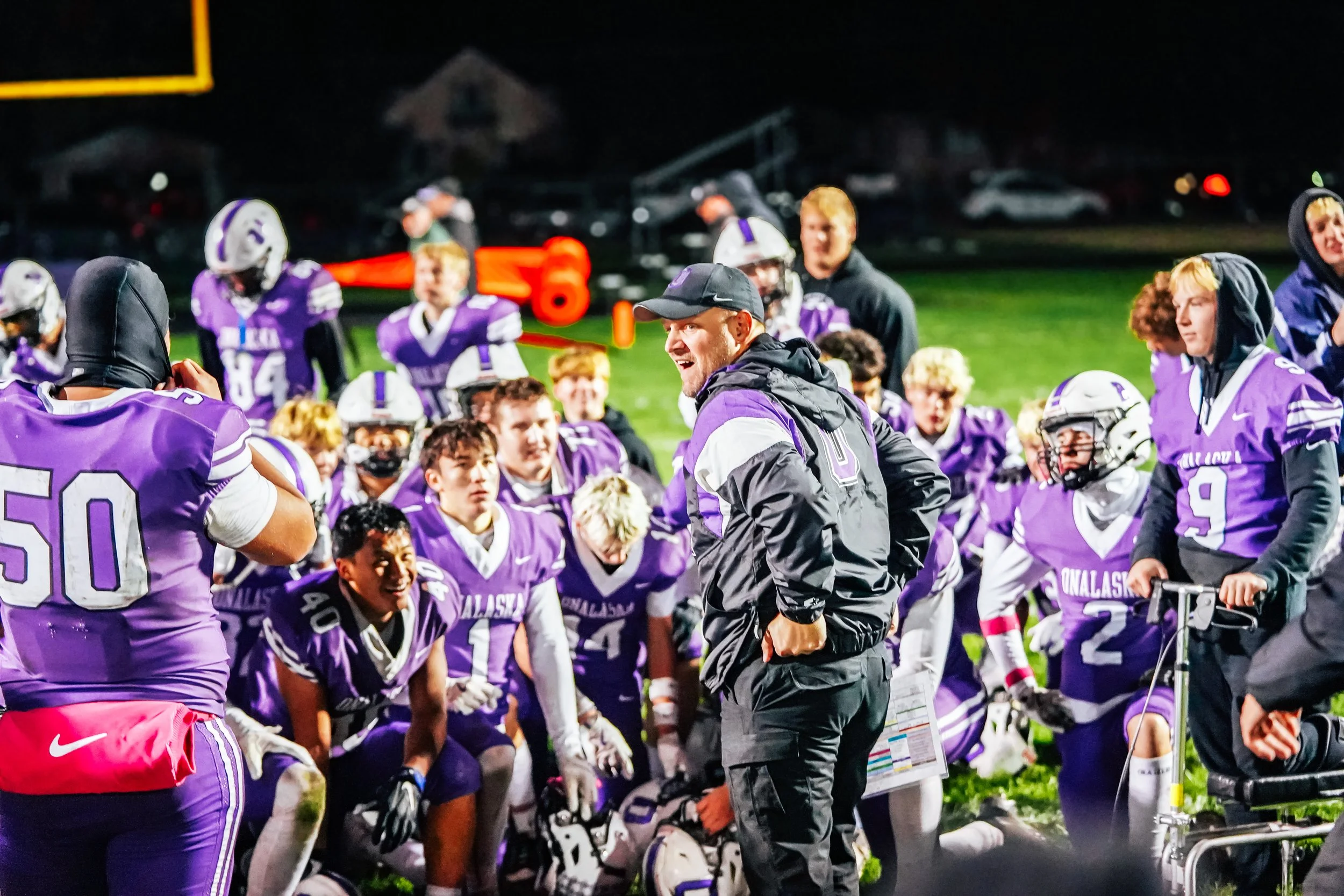 Football team wearing purple and white uniforms gathered around their coach during a timeout on a football field at night.
