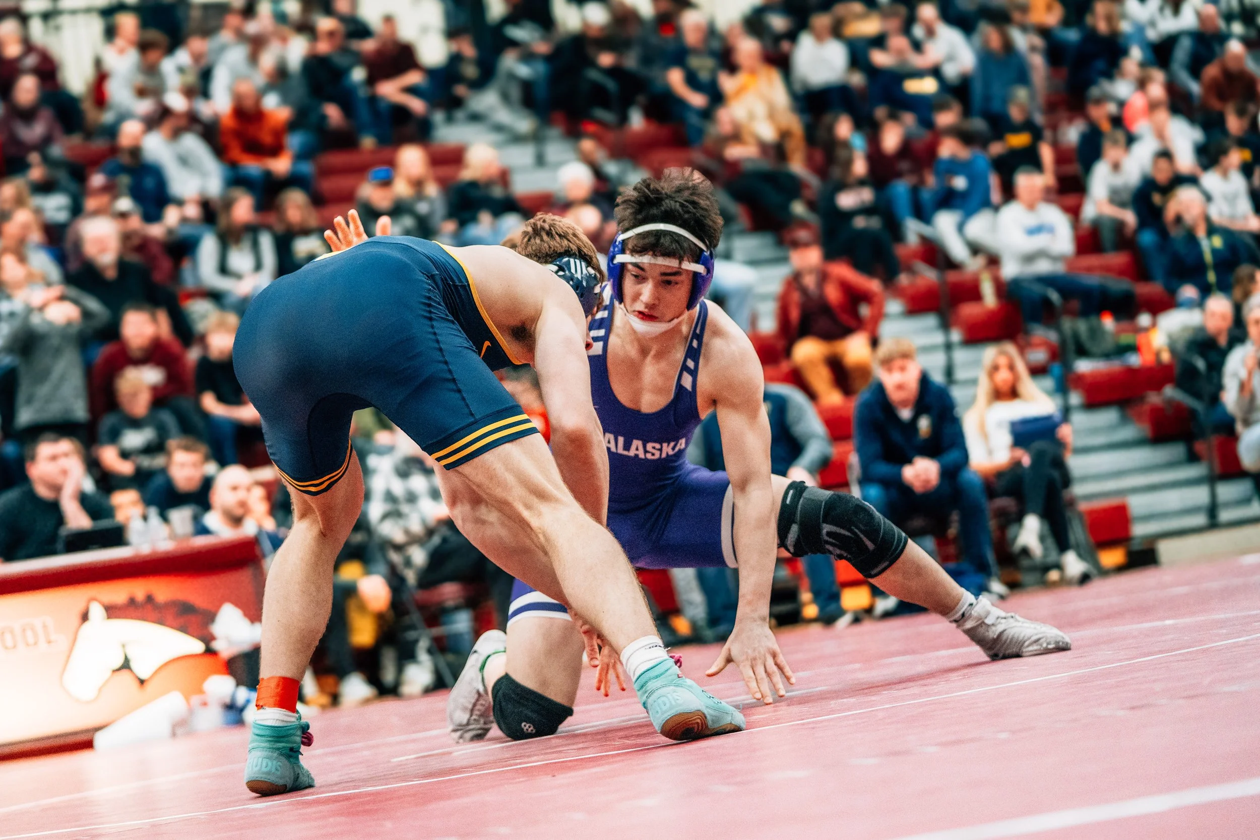 Two wrestlers in singlets engaged in a match on a wrestling mat, with spectators watching in the background.