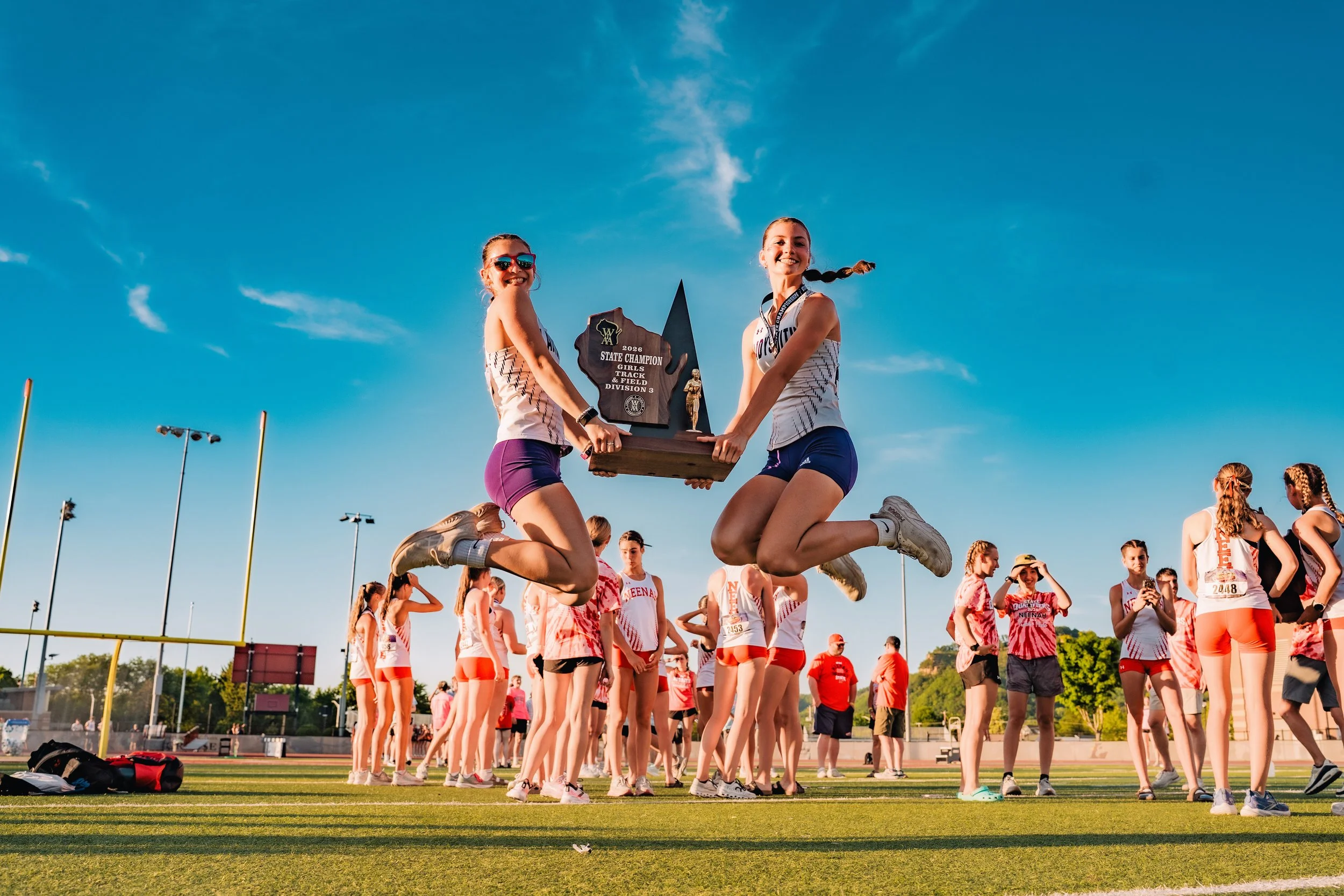 Two young women in athletic attire are jumping in the air and holding a large trophy with a wooden plaque that reads "2026 State Champion Girls Track & Field Division 3" on a sports field. They are surrounded by other athletes, some standing, with a 