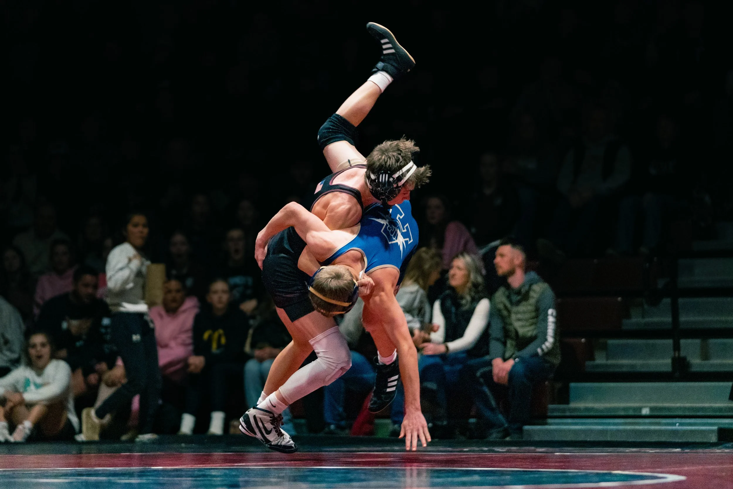 Two wrestlers in a wrestling match, one in a black singlet and the other in a blue singlet, with the wrestler in black lifting the wrestler in blue off the ground during a match with spectators watching.