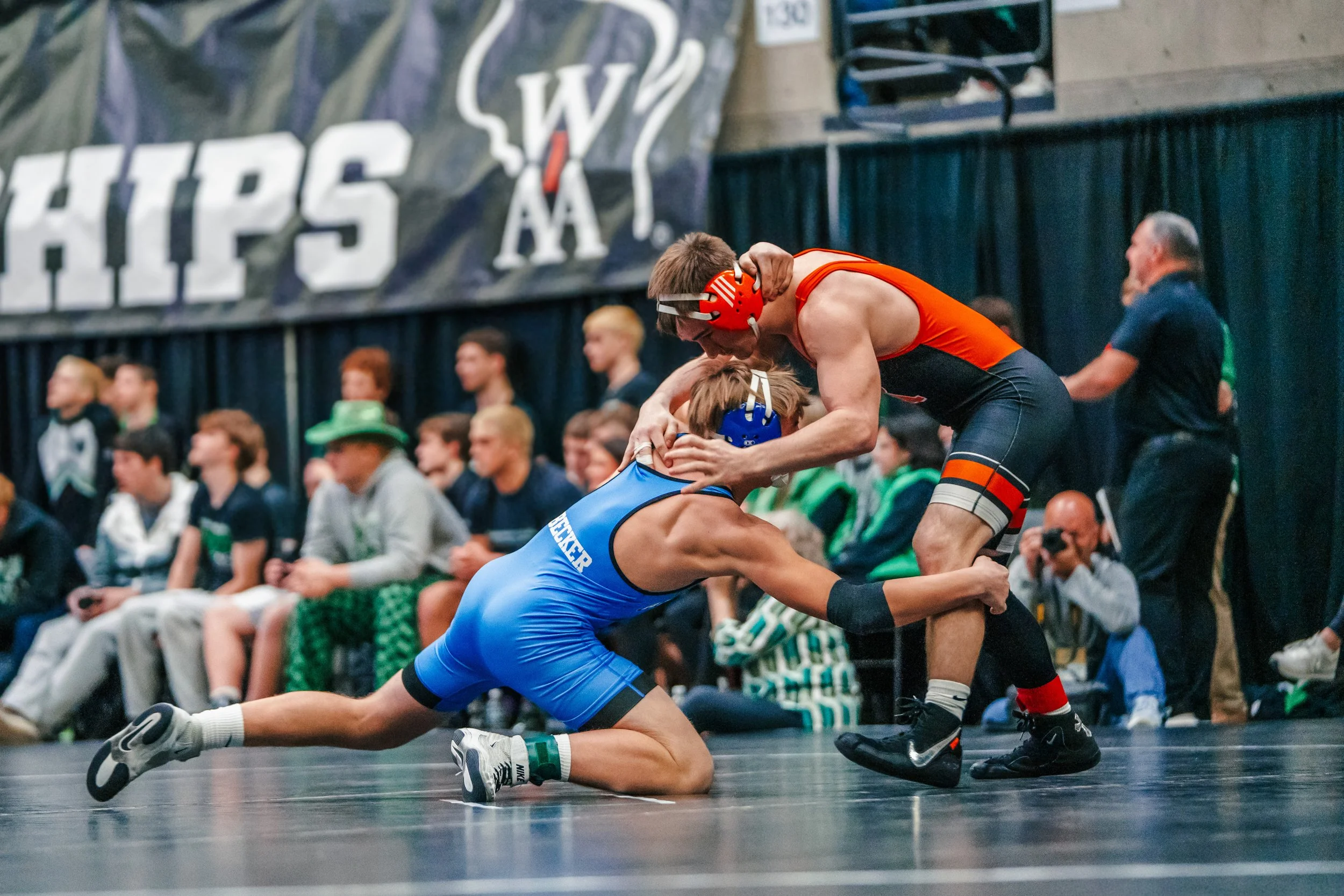 Two male wrestlers with athletic builds competing on a wrestling mat, with one in a blue singlet on the ground and the other in a red and black singlet standing over him. Spectators are seated in the background at an indoor wrestling event.