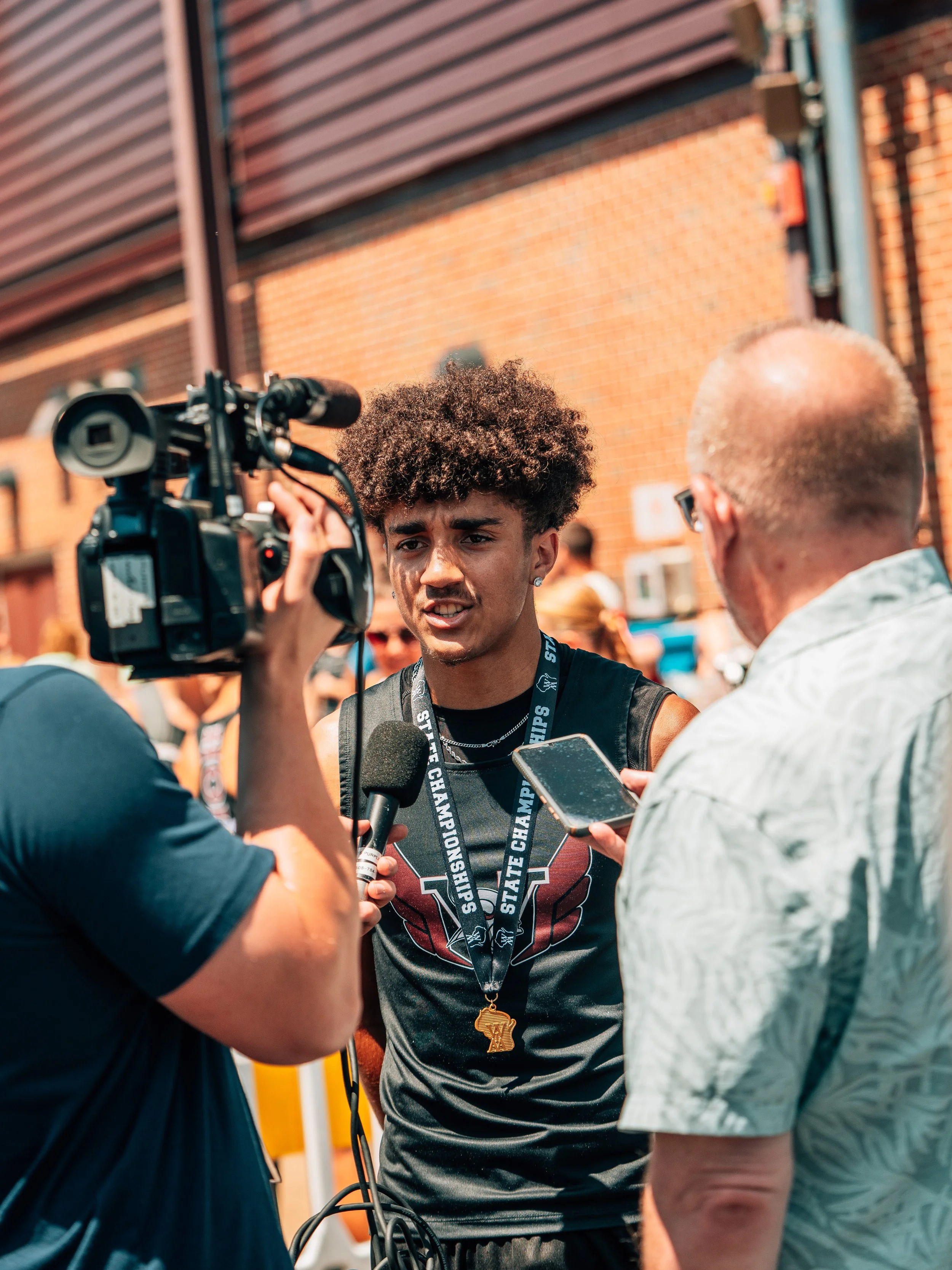 A young man wearing a medal and lanyard with 'State Champions' text is being interviewed outdoors by a group of reporters. The scene is set against a brick wall and sunlight.