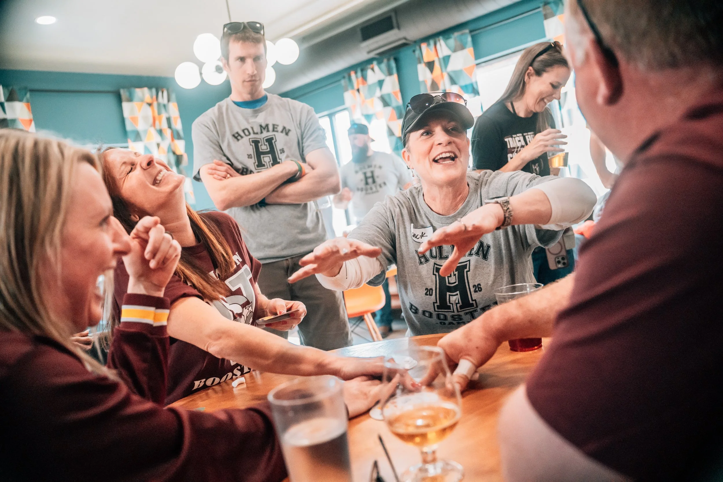 Group of people, some wearing Harvard logos, sitting around a table laughing and engaging in conversation at a social gathering or party.
