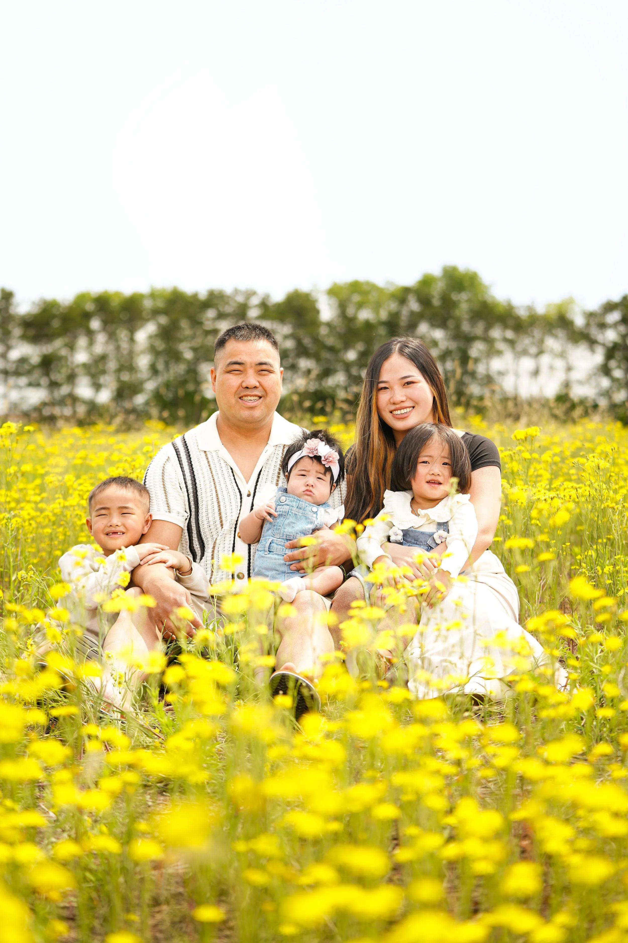 A family of six, including two adults and four children, sitting in a yellow flower field outdoors against a background of green trees and a bright sky.