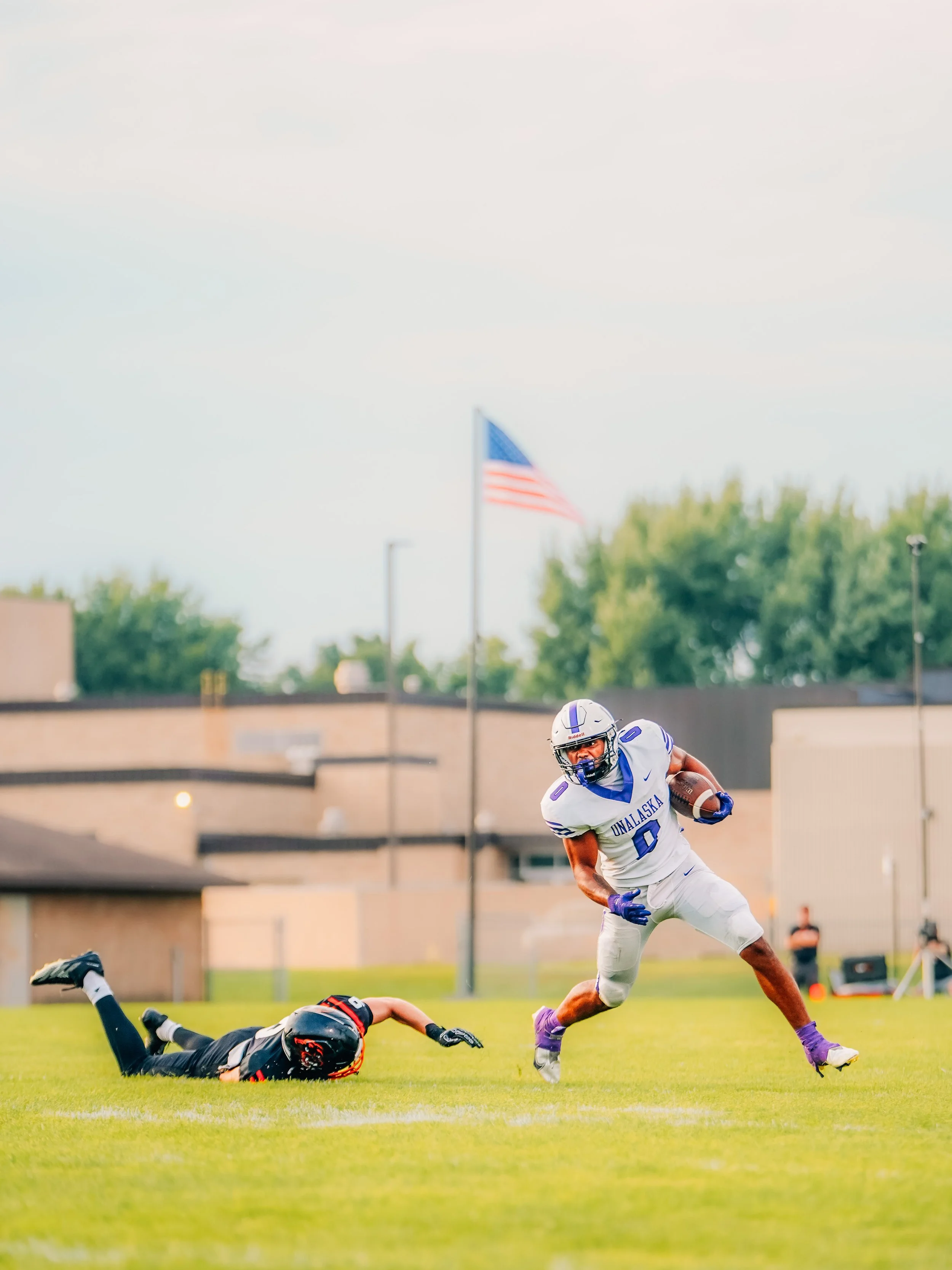 A football player wearing a white uniform with purple details and the number 0, running with the ball as a player in a black uniform dives to the ground.