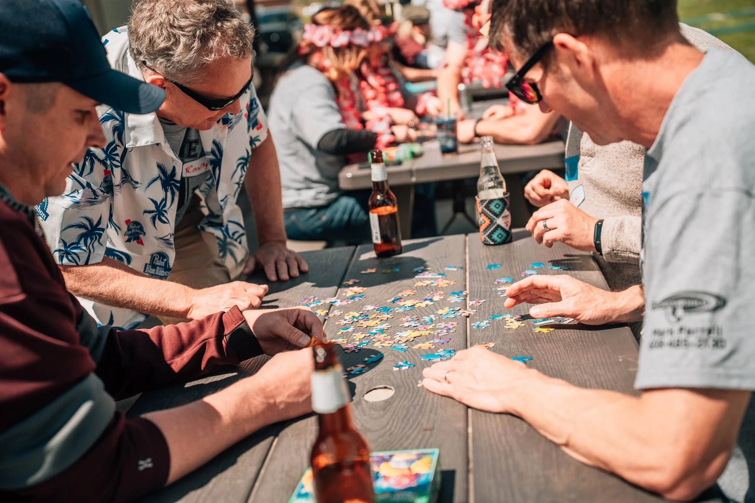 Group of people playing jigsaw puzzle at outdoor social gathering with tables, drinks, and background of people and trees.