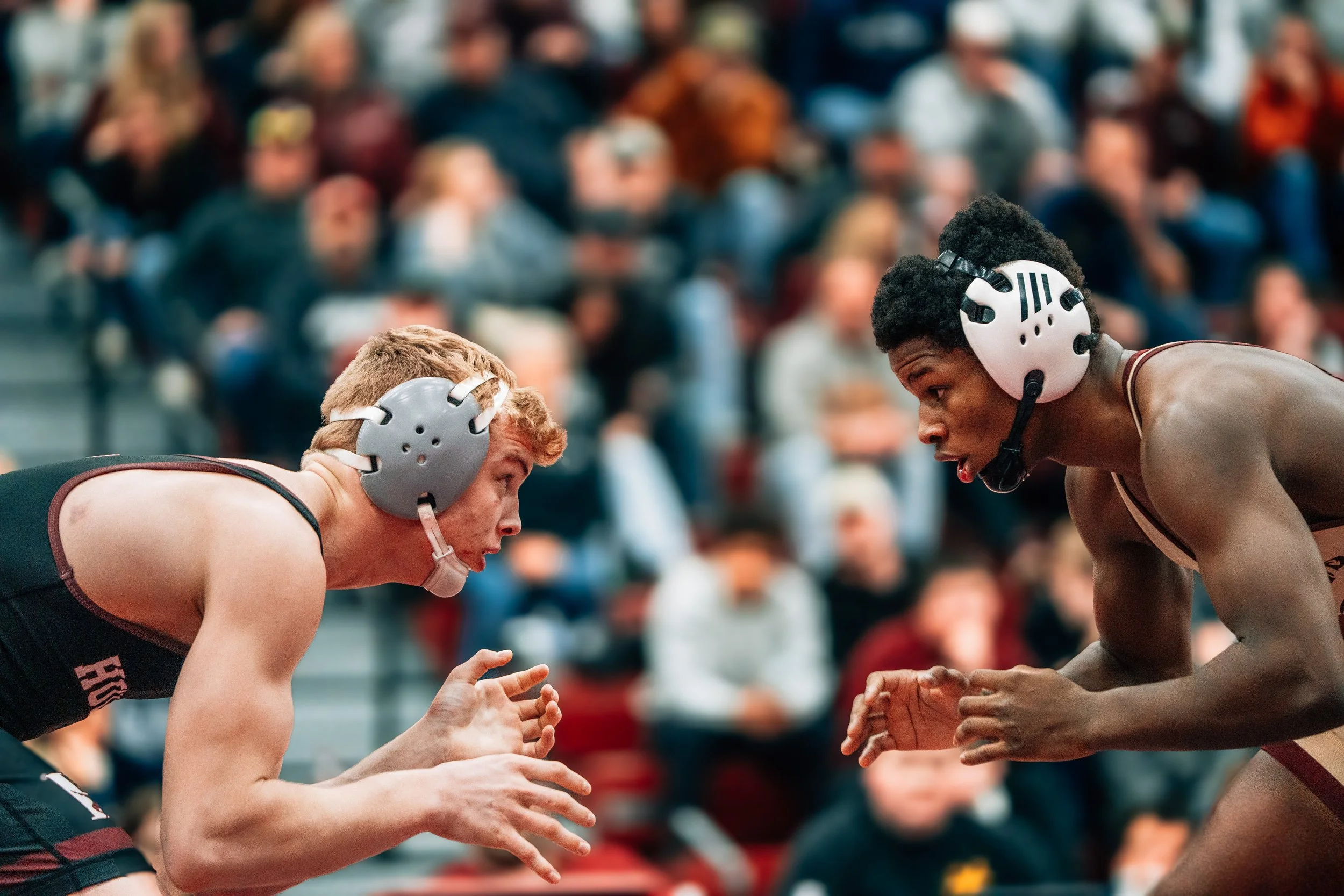 Two wrestlers face each other in a match, wearing wrestling gear and helmets, with a crowd of spectators watching in the background.