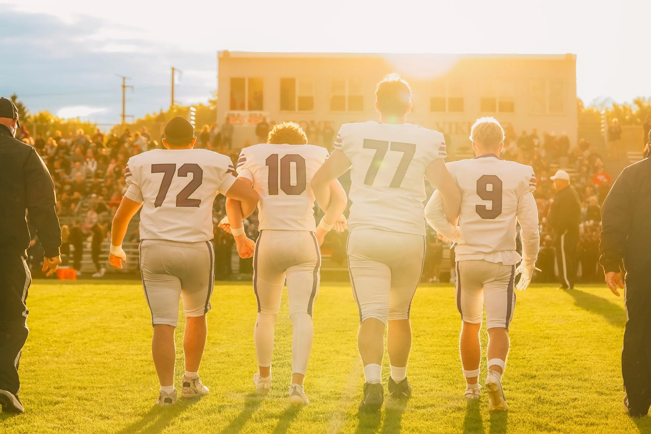 American football players walking onto the field during a game in the late afternoon or early evening with sunlight behind them, with cheerleaders and spectators in the background.