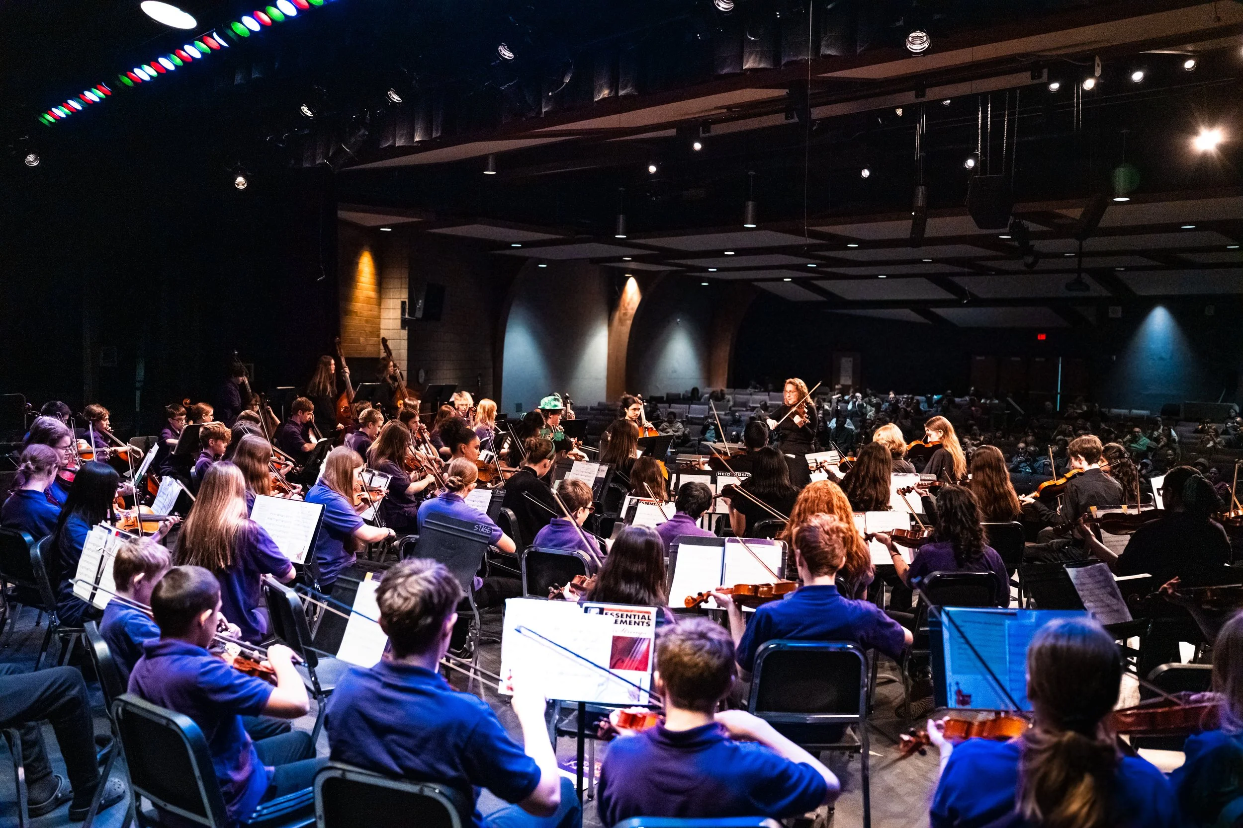 An orchestra performing on stage in a concert hall, with musicians playing violins, cellos, and other instruments, directed by a conductor, with audience seating visible in the background.