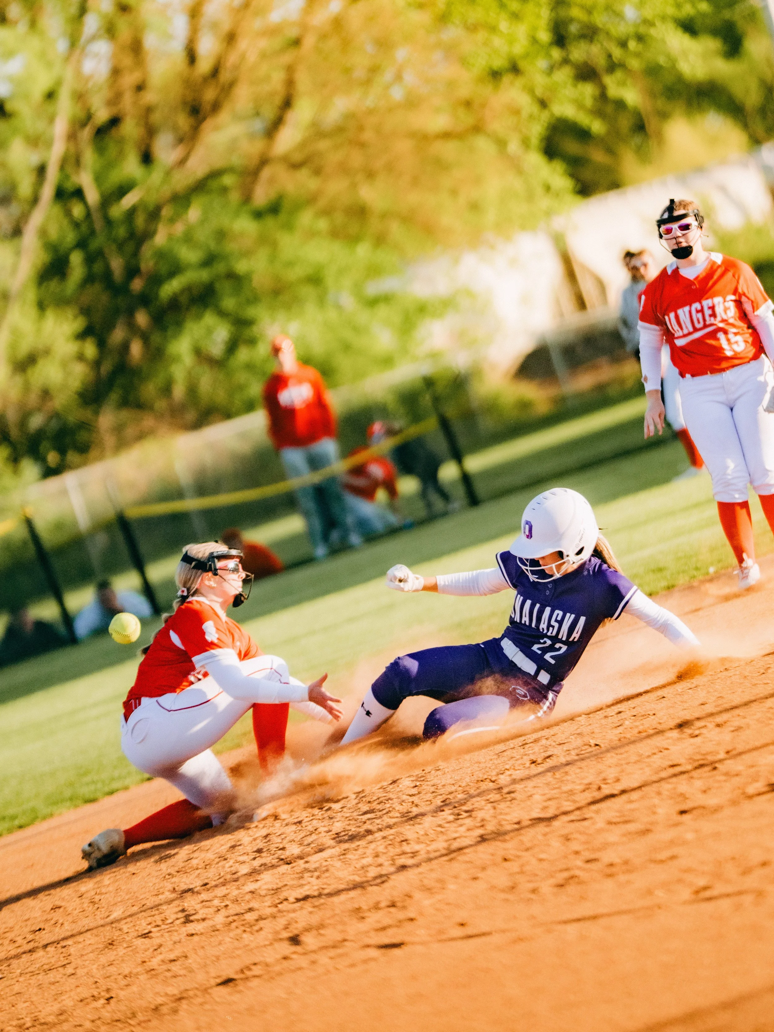 A softball game in progress with players sliding into a base, including a player from Alaska in navy, another in red, and a player in red attempting to catch a ball during a slide on the dirt field, with other players and spectators in the background
