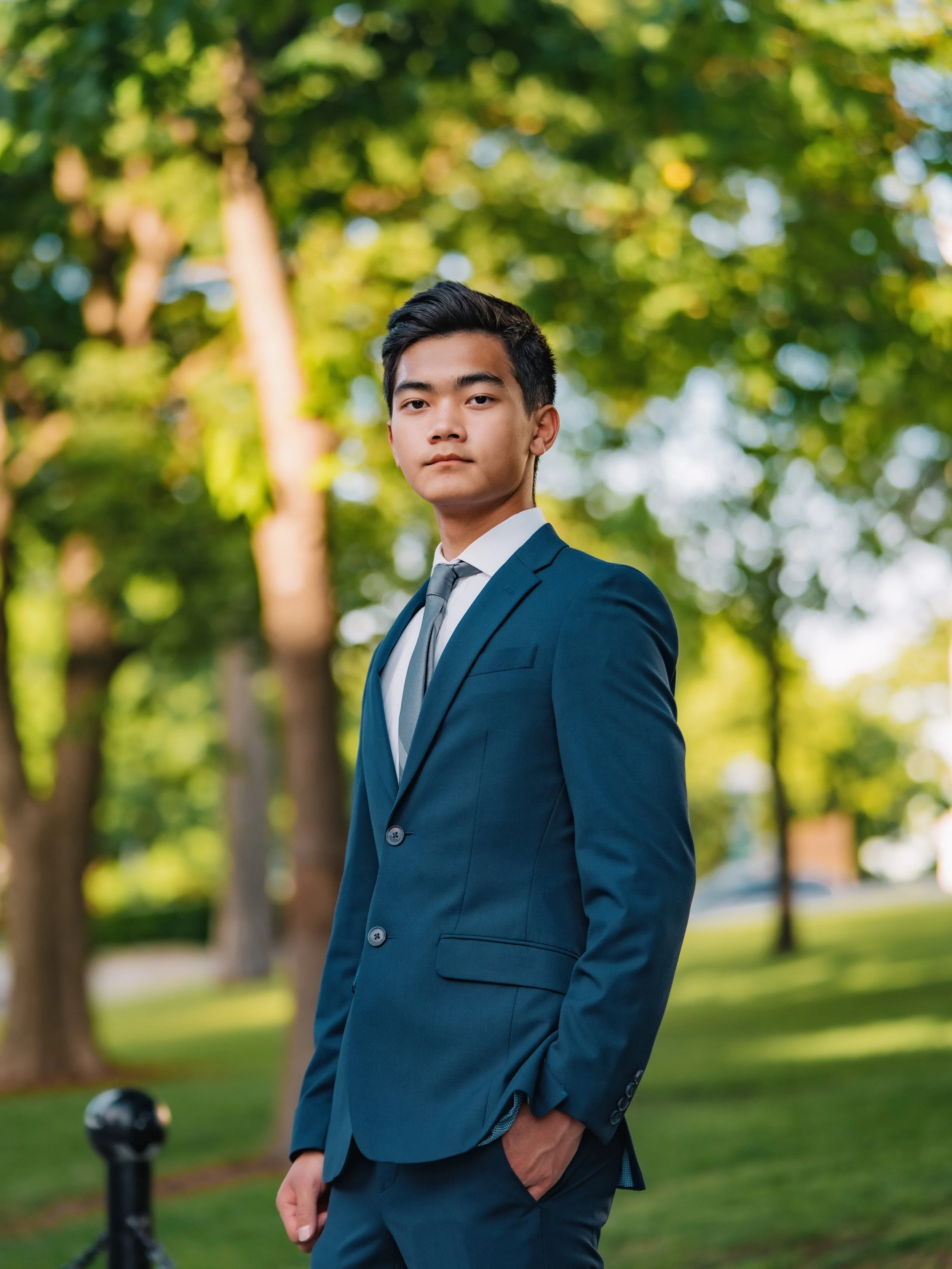 A young man dressed in a blue suit and tie stands outdoors in a park with trees and greenery in the background. He is looking confidently at the camera with one hand in his pocket.