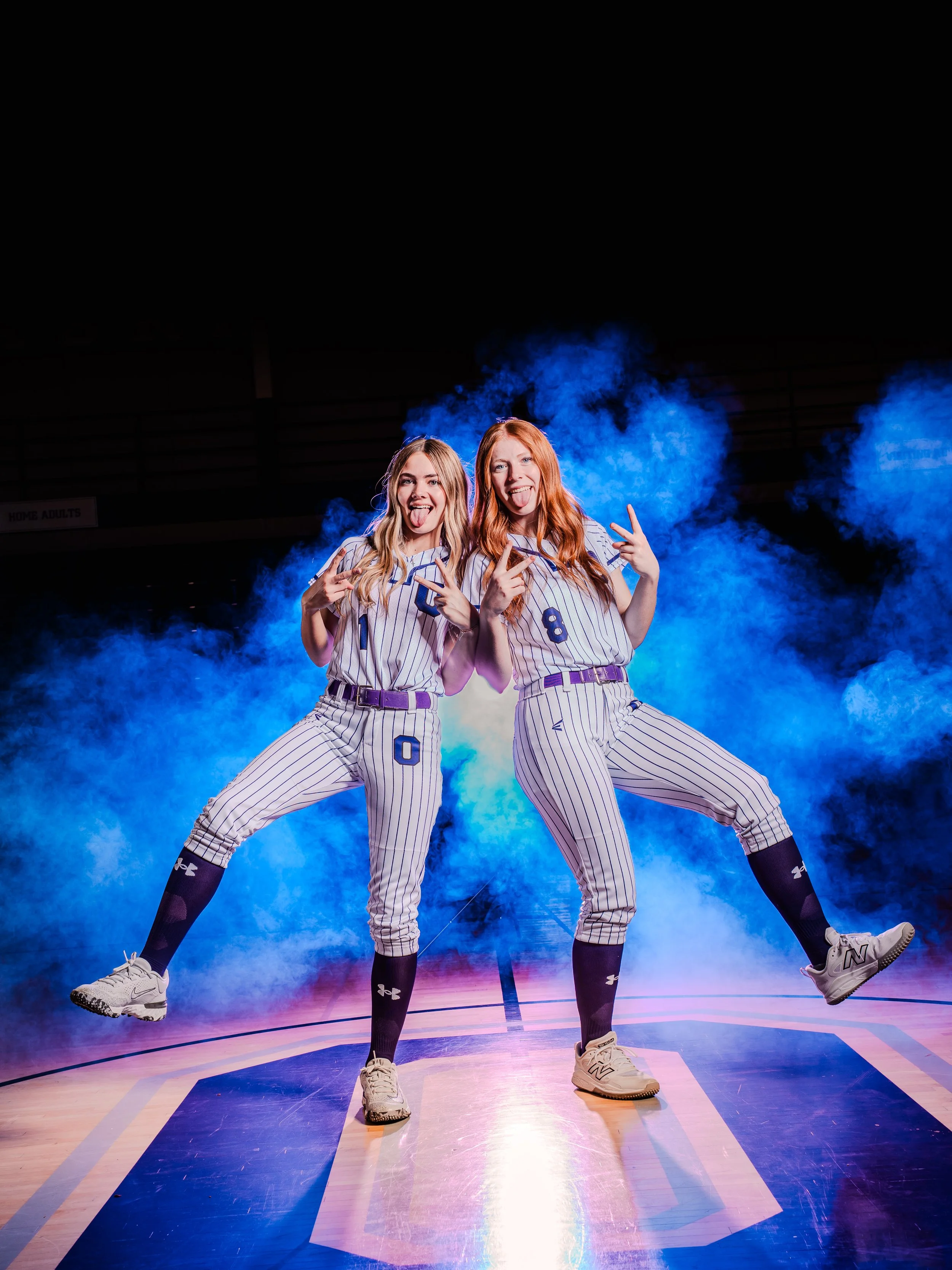 Two young women in baseball uniforms dancing on a basketball court with blue smoke in the background.