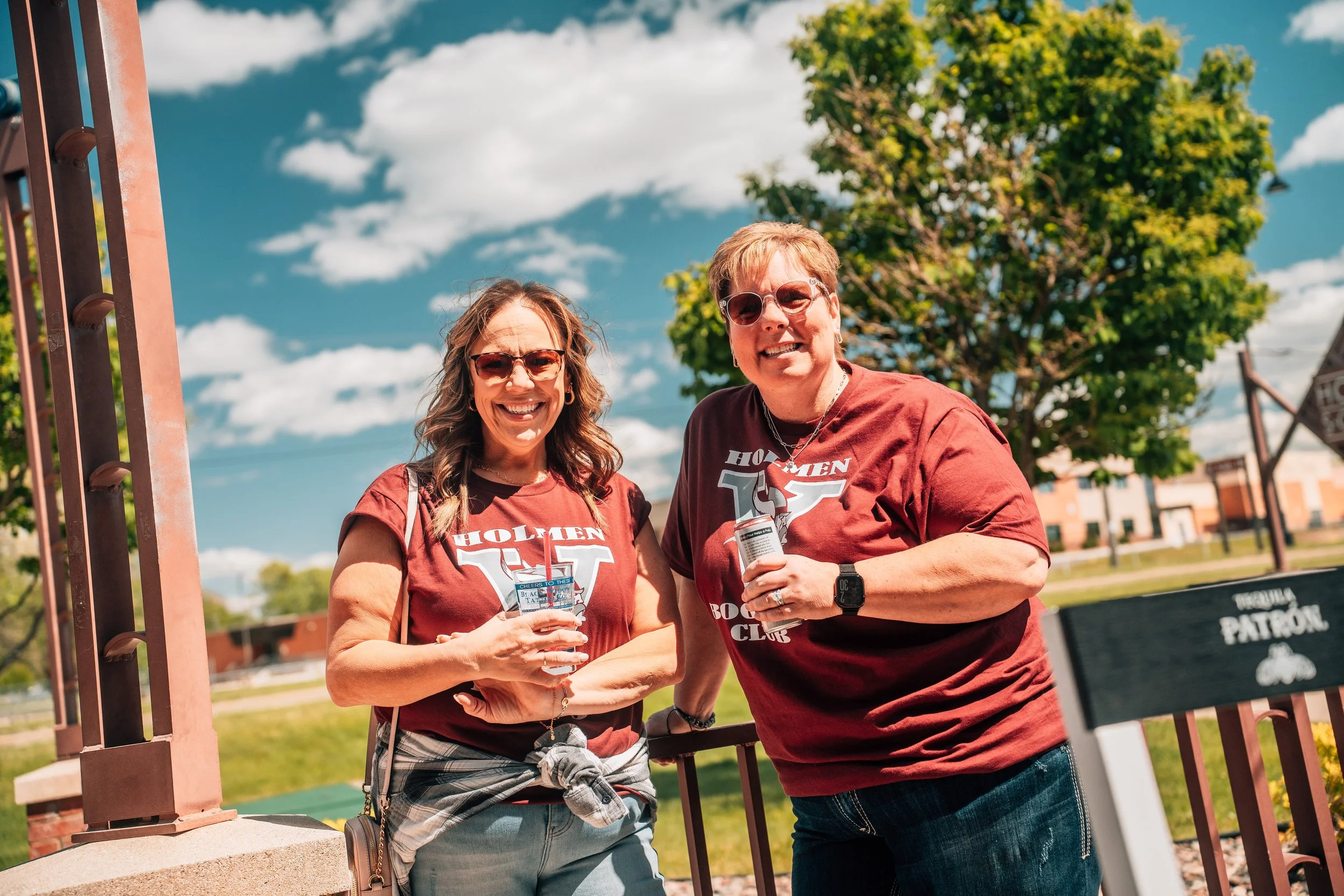 Two smiling women wearing maroon 'Holmen' T-shirts standing outdoors under a blue sky with clouds, near a pink metal structure and a green tree.