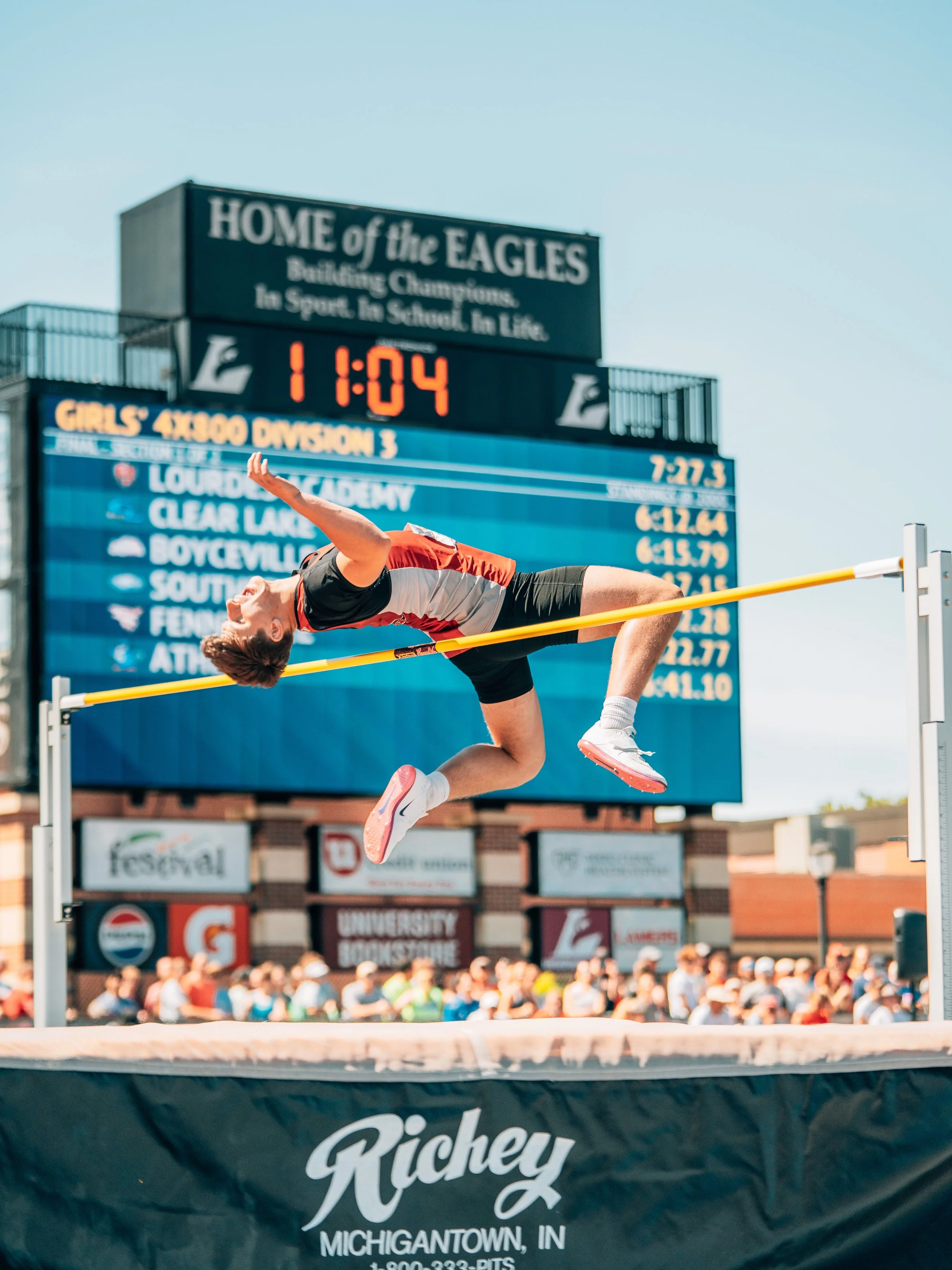 A male athlete performing a high jump at a track and field stadium, with a crowd of spectators and a scoreboard in the background showing various race times and team names.
