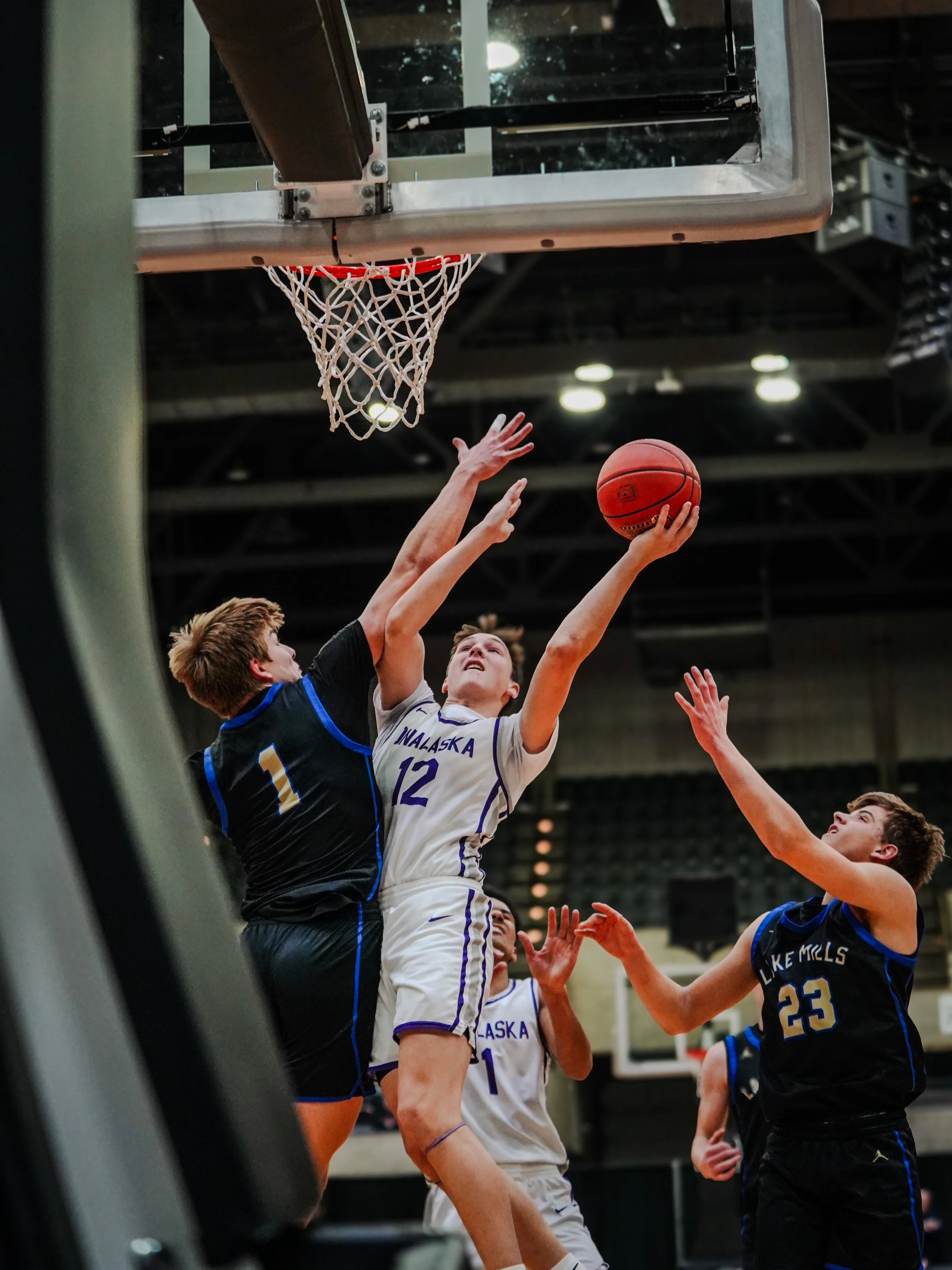 Two basketball players are competing near the hoop. One player in a white jersey with number 12 from Alaska is jumping to shoot or block, while a player in a black jersey with number 23 from Lake Mills is reaching to block. The scene is inside an ind