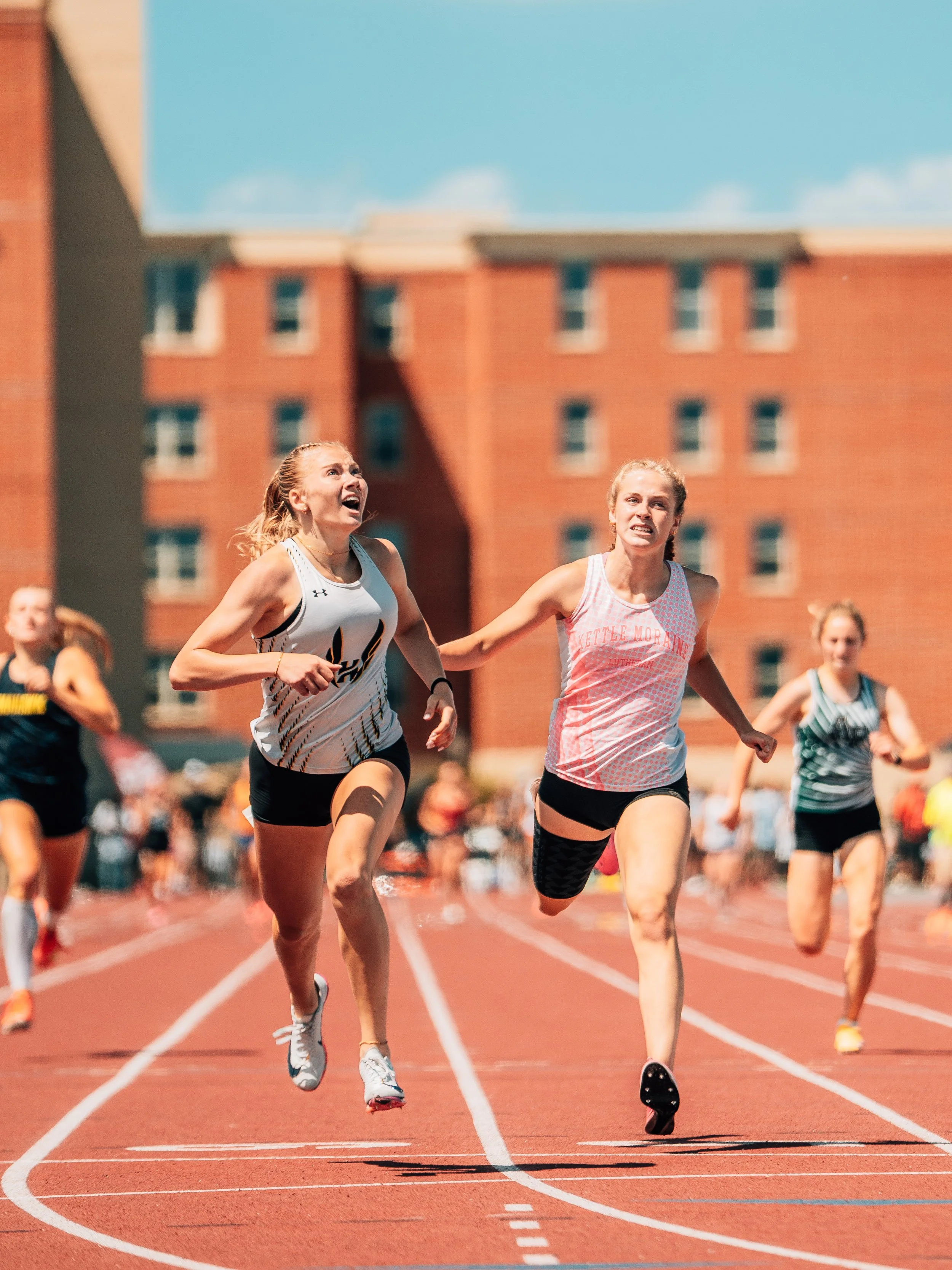 Two female athletes competing in a race on a red running track, with a brick building and a clear blue sky in the background.