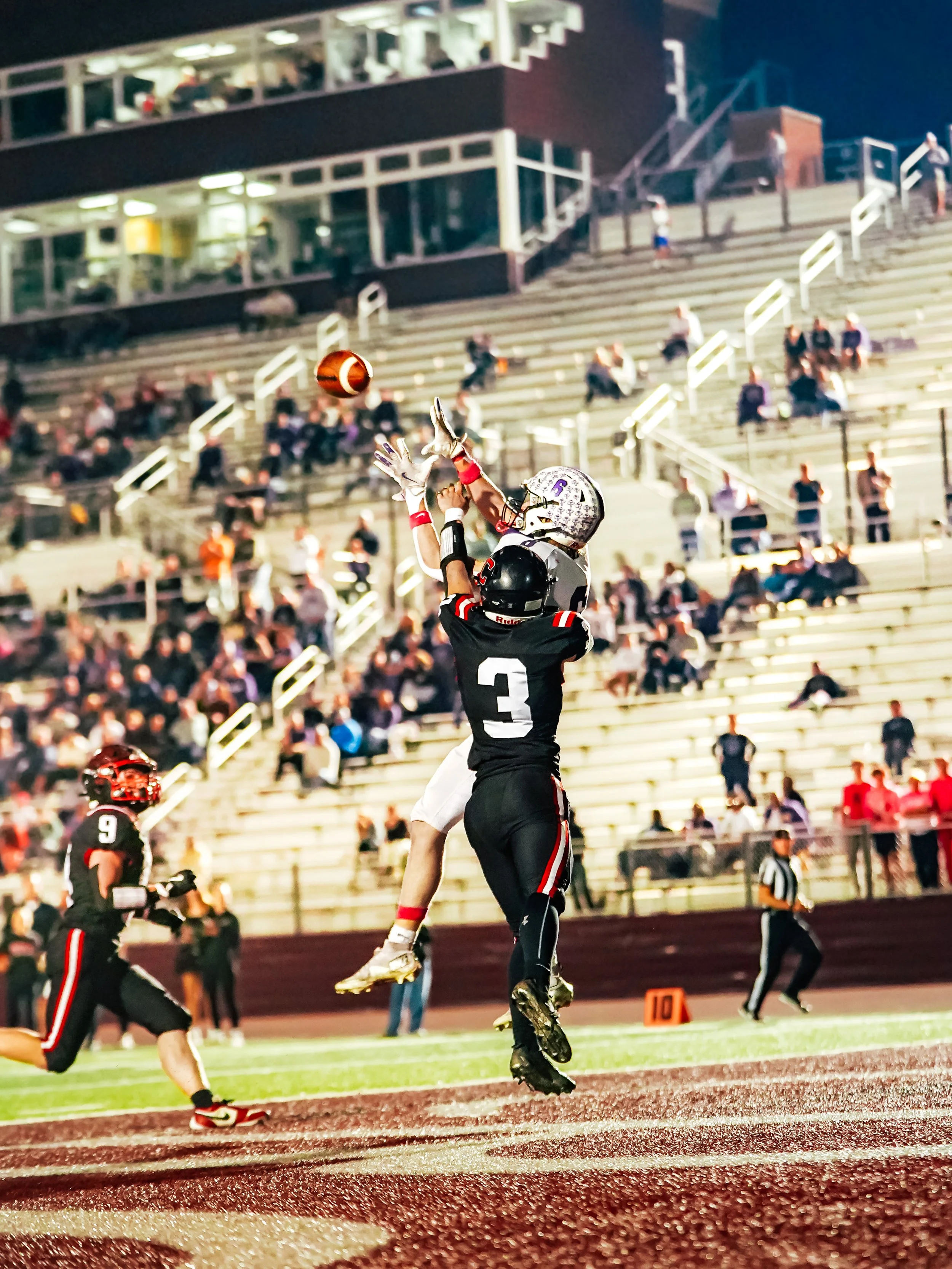 A football player in black and red uniform jumping to catch the ball during a game at night, with other players and spectators in the background.