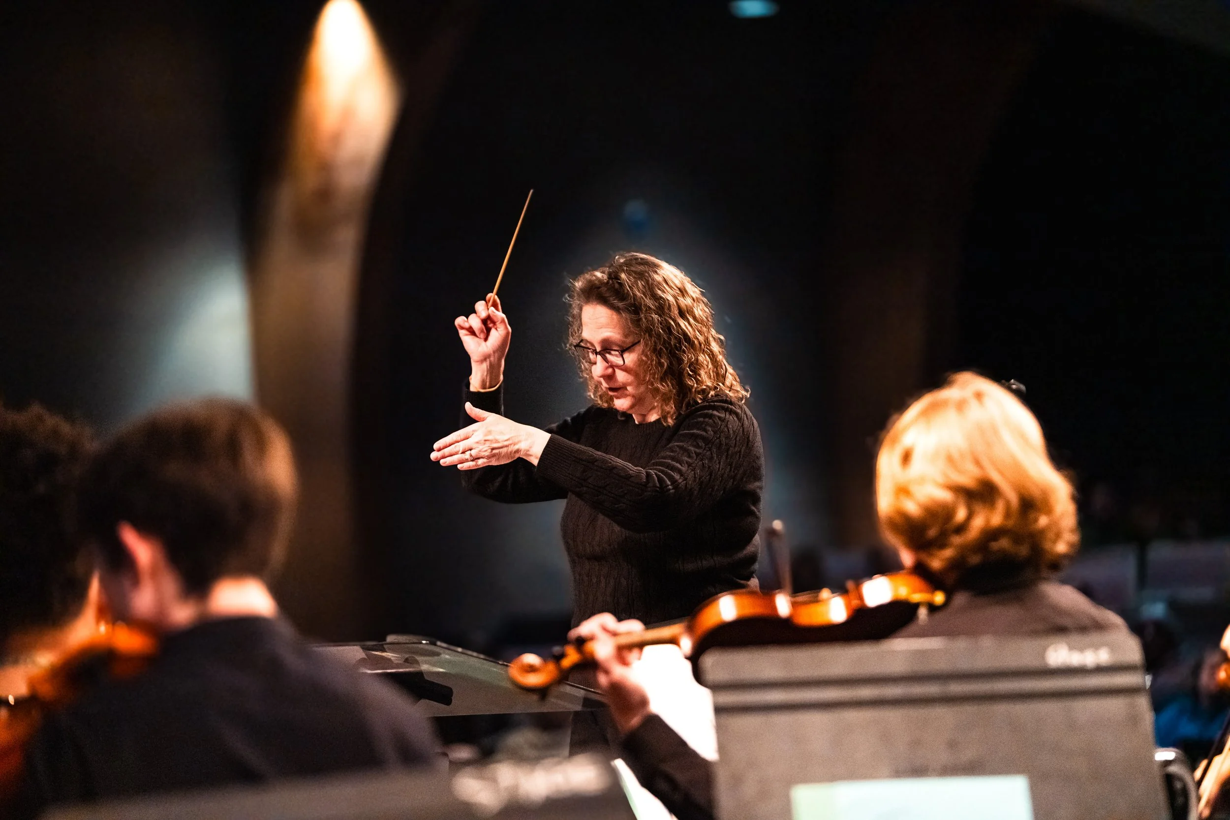 A woman conducting an orchestra, holding a baton, with musicians playing violins in the foreground.