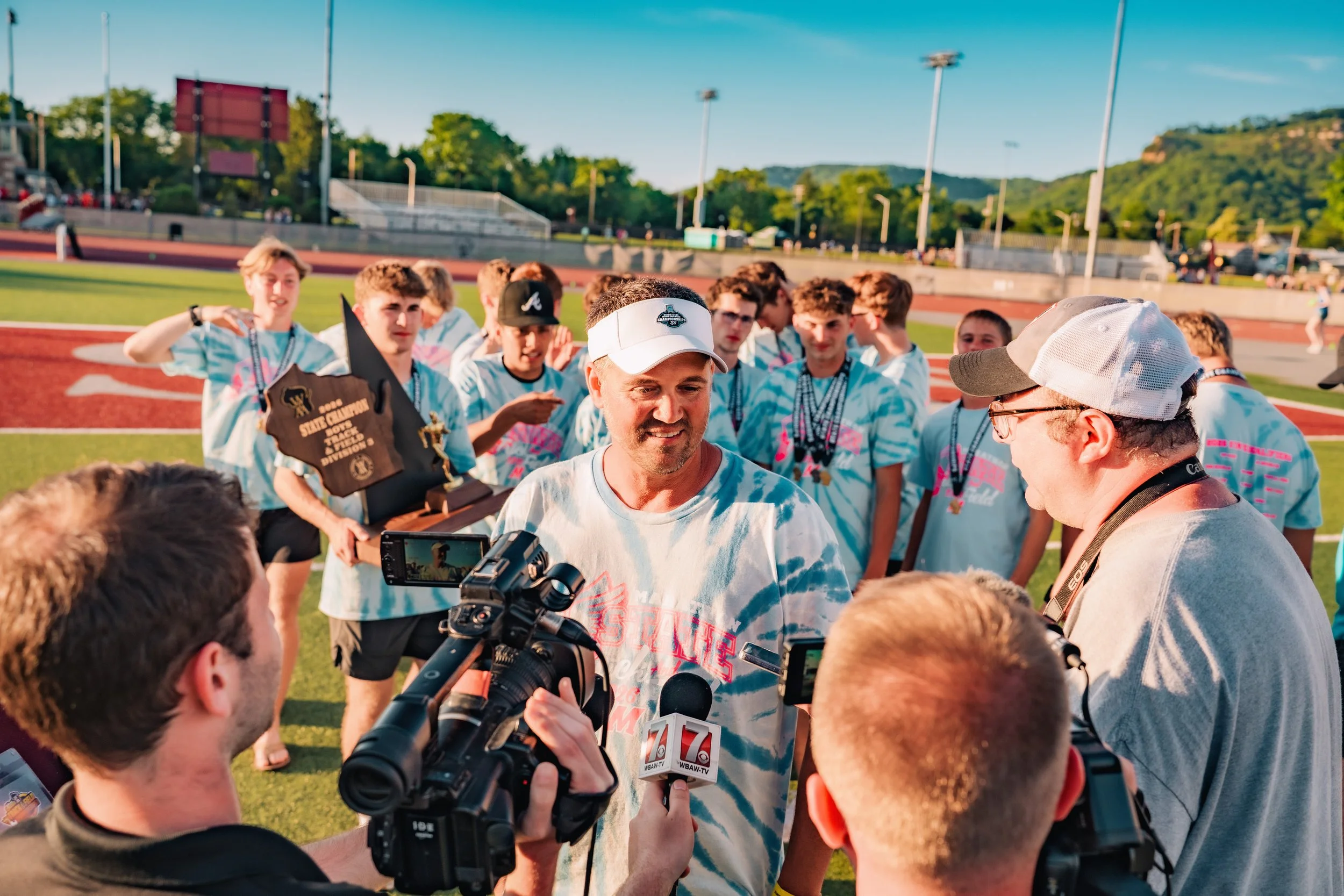 A group of young athletes in tie-dye shirts standing on a track field, being interviewed by media, with hills and a bright sky in the background.
