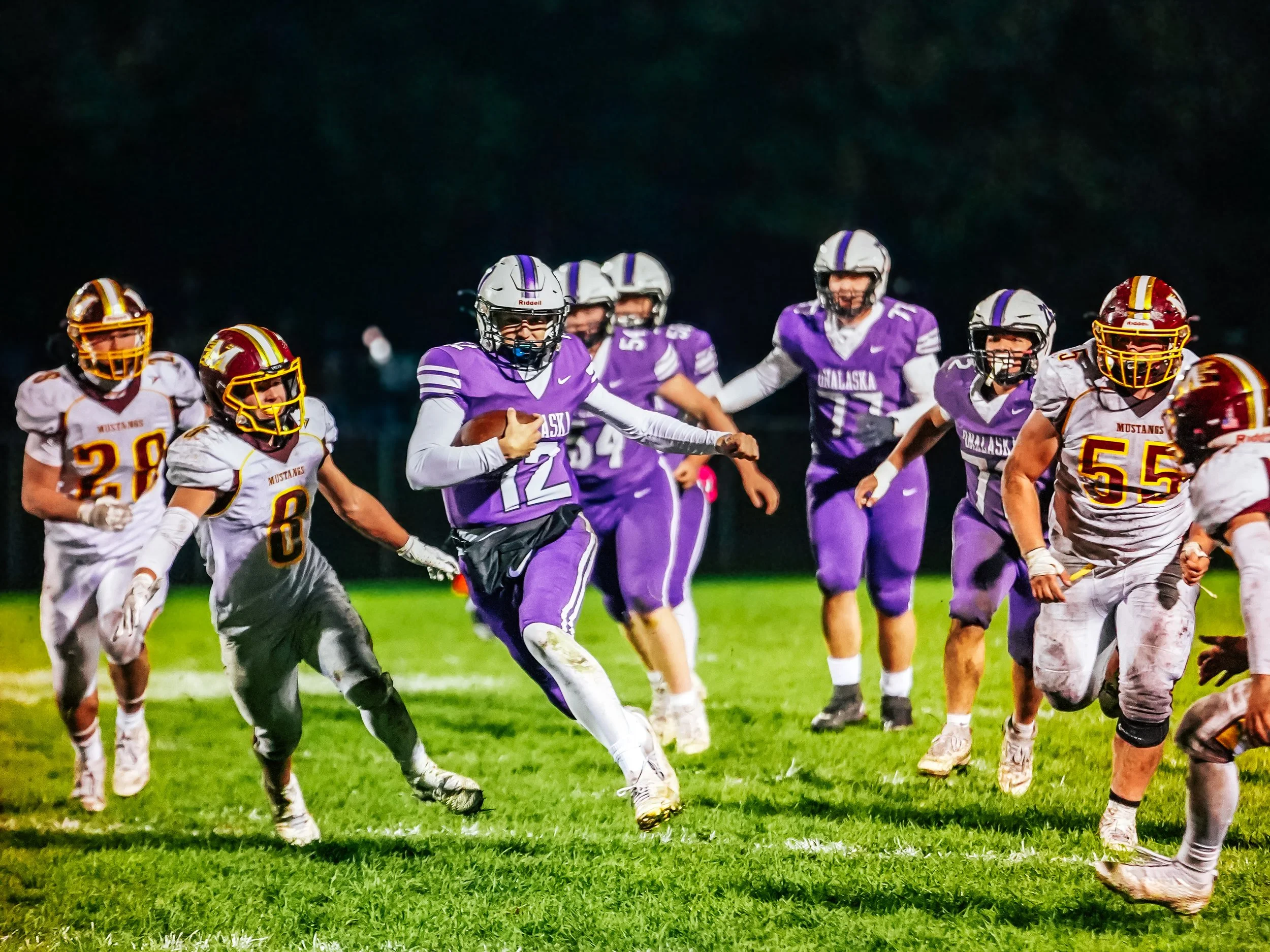A group of American football players wearing purple and white uniforms from Alaska and white and maroon uniforms from Mustang are on a field during a game at night, with players running and some holding a football.