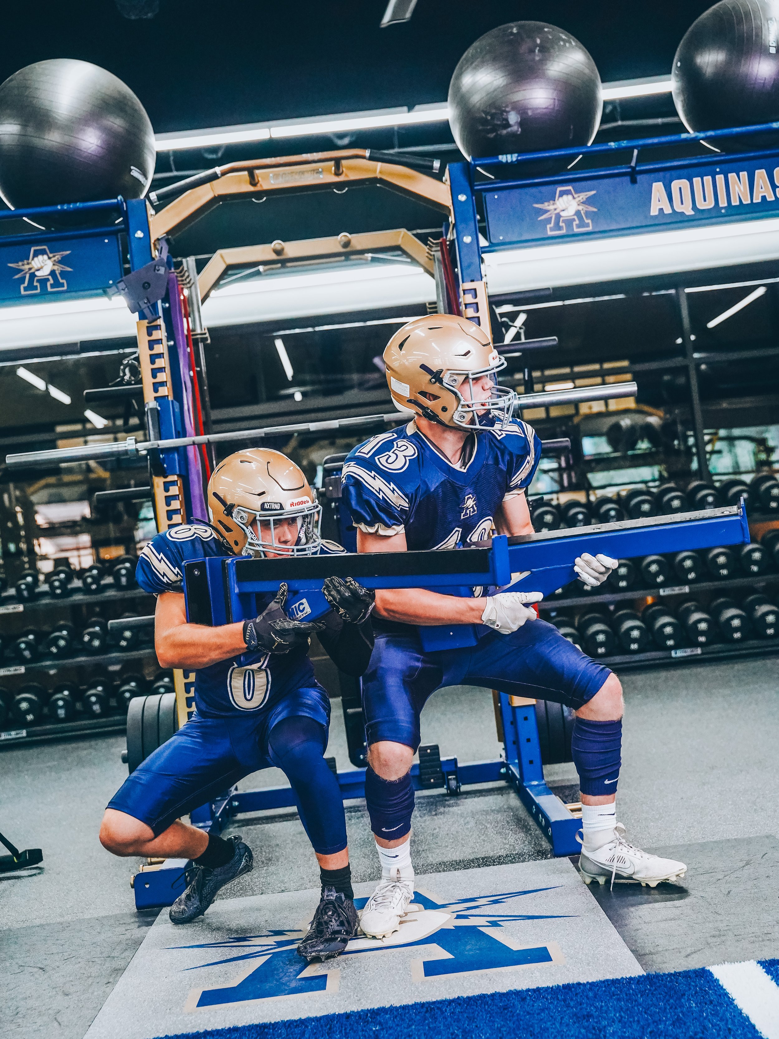 Two football players working out with a weightlifting machine in a gym, wearing blue uniforms and gold helmets, with gym equipment and dumbbells in the background.