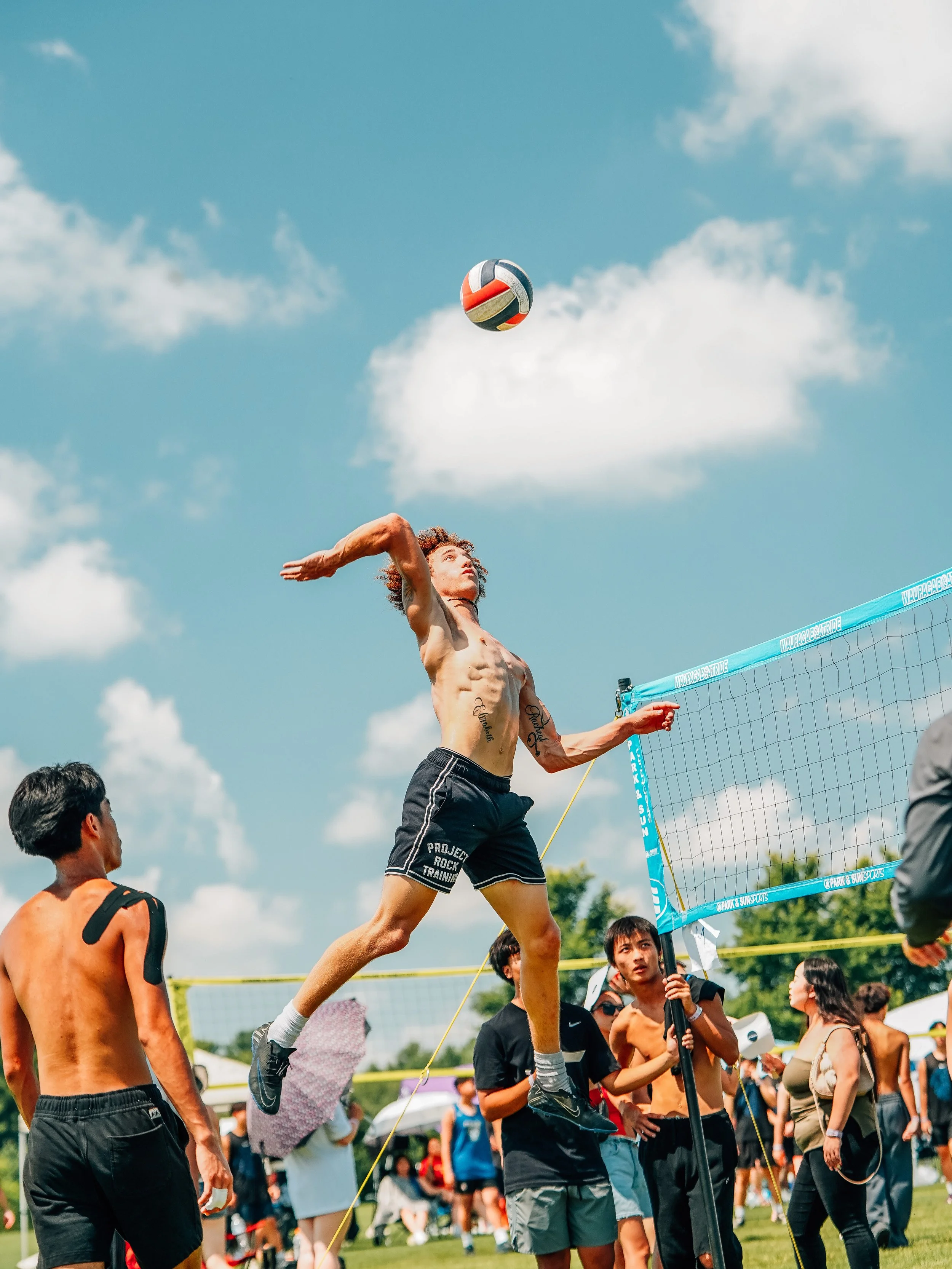 A man jumps high to hit a volleyball over a net during an outdoor game, with other players and spectators around under a blue sky with clouds.