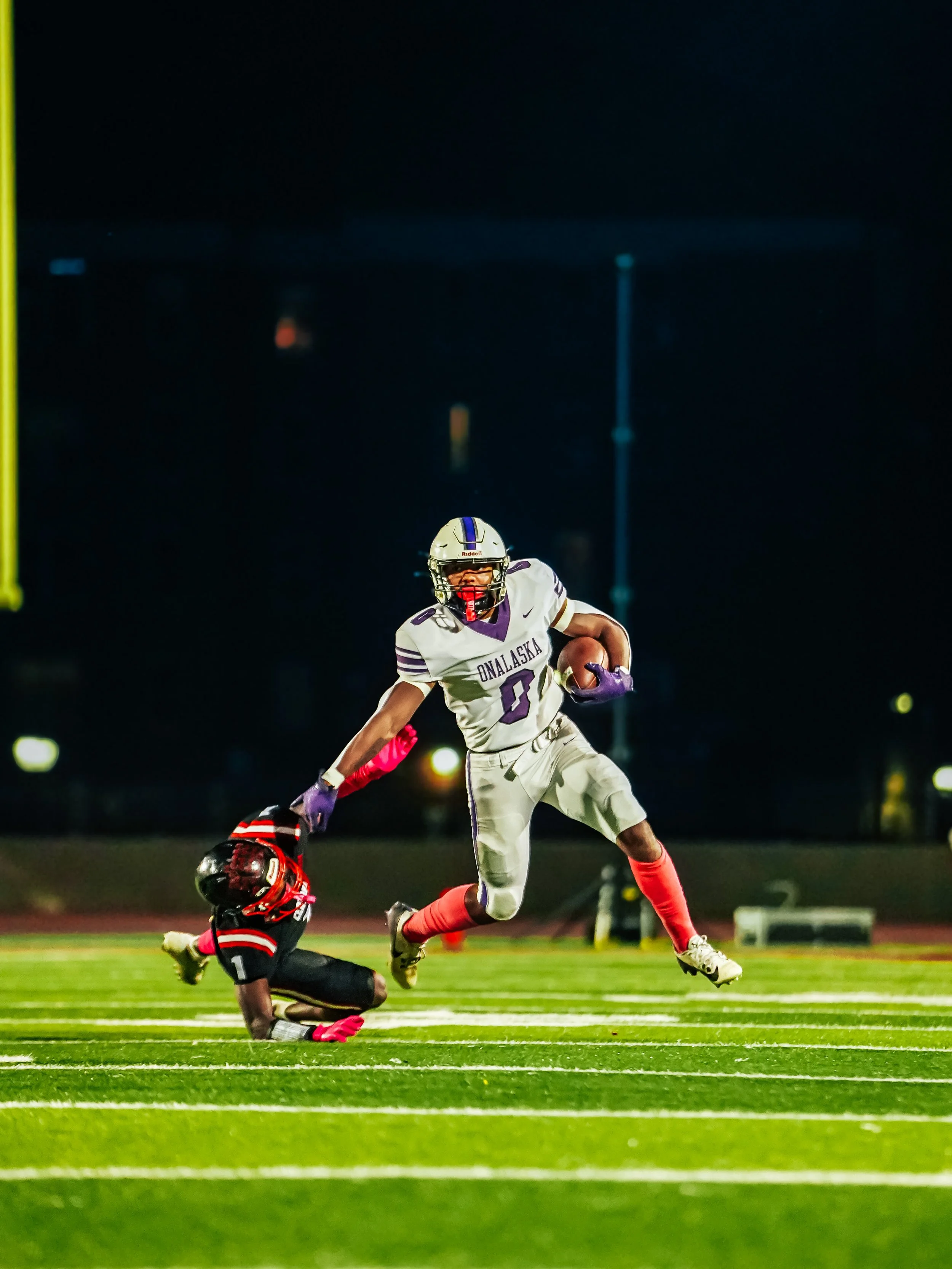 A football player in a white and purple uniform running with the ball past a fallen opponent in a black and red uniform on a brightly lit football field at night.