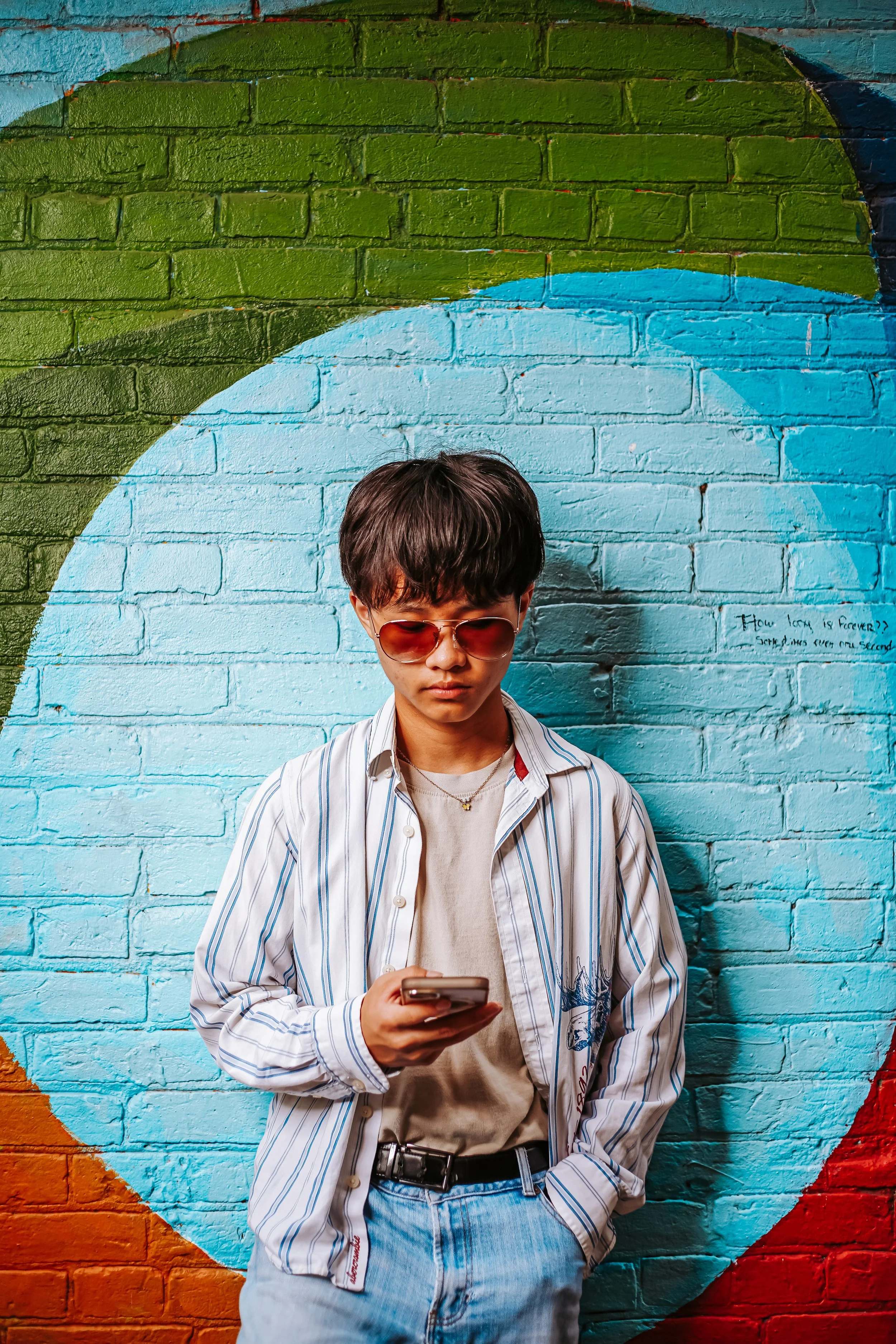 A young man with dark hair wearing sunglasses, a striped shirt, and light jeans stands in front of a colorful mural featuring a large design on a brick wall. He is looking at his phone, with his face partially visible.
