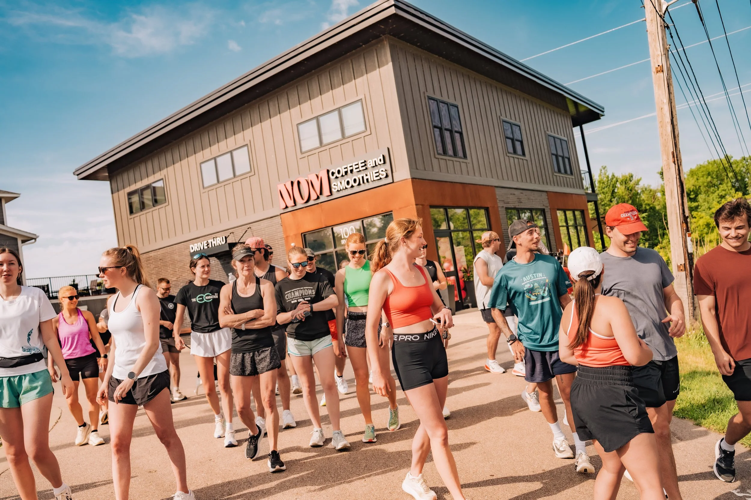 A group of people walking outside on a sunny day, with a building in the background that has a sign reading 'MOM Coffee and Smoothies.'