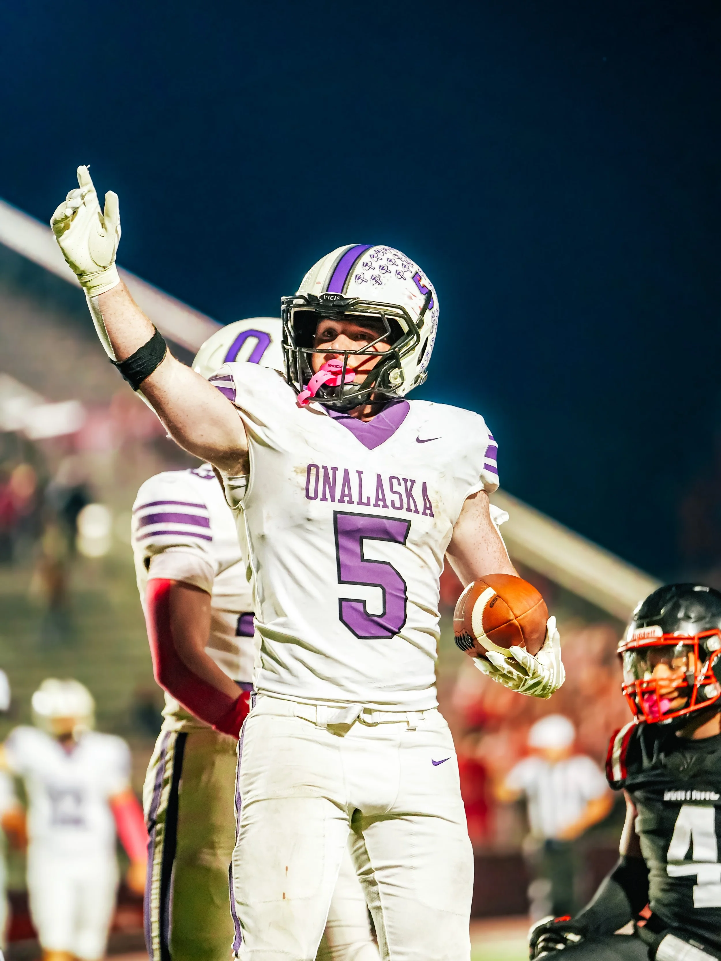 American football player in white and purple uniform holding a football, pointing with his right hand, wearing a helmet with purple and white design, during a game.
