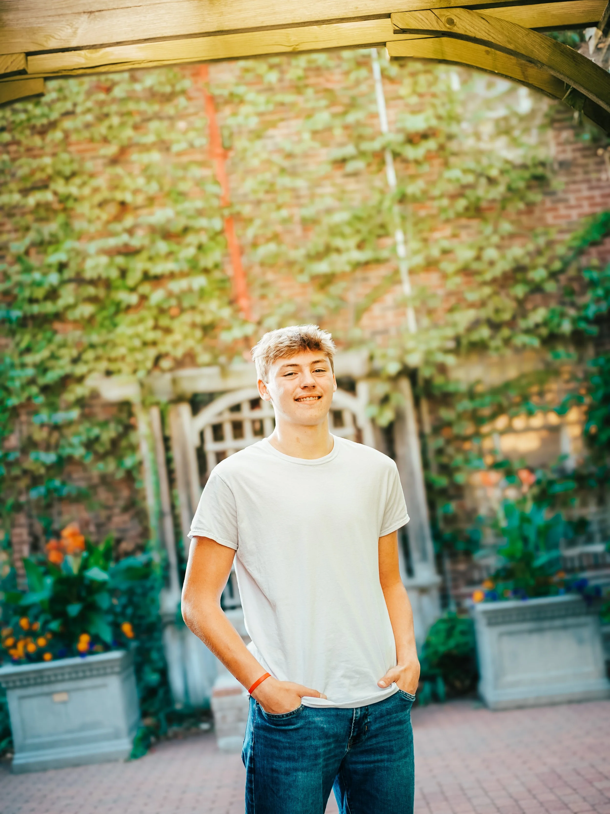 Teenage boy standing outdoors in front of a brick wall and a garden with plants and flowers, smiling at the camera.